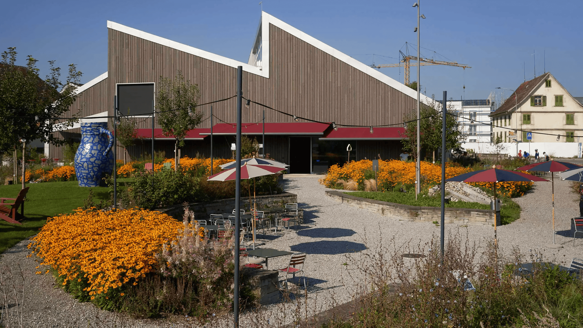 Ein modernes Gebäude mit einem Zickzack-Dach ist umgeben von leuchtend gelben Blumen, Sitzgelegenheiten im Freien mit Sonnenschirmen und Kieswegen an einem sonnigen Tag. In der Nähe des Eingangs steht eine große blau gemusterte Skulptur.