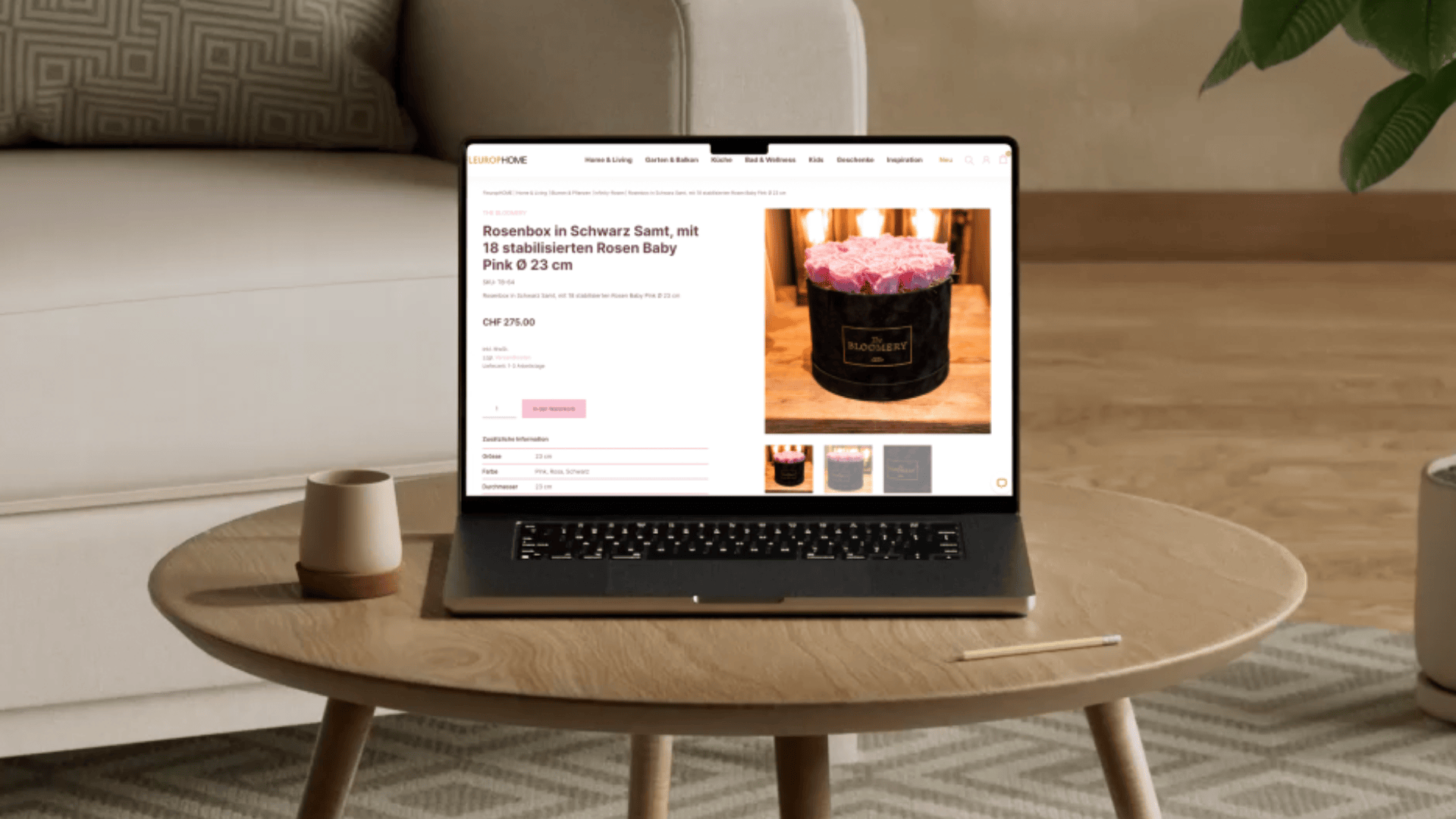 Ein Laptop auf einem Couchtisch zeigt eine Website an, auf der eine Rosenbox aus schwarzem Samt mit rosa Rosen verkauft wird. Auf dem Tisch stehen auch ein beiger Becher und ein Stift. Im Hintergrund stehen ein beiger Sessel und eine grüne Pflanze.
