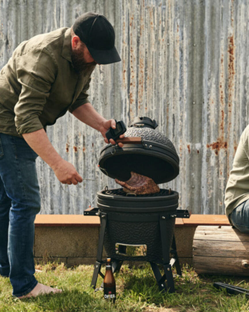 Ein Mann öffnet den Deckel eines schwarzen Keramikgrills, um zu prüfen, ob das Fleisch im Inneren brutzelt, draußen im Gras, mit einer Wellblechwand und einer Bierflasche in der Nähe. Eine andere Person sitzt teilweise außerhalb des Bildes.