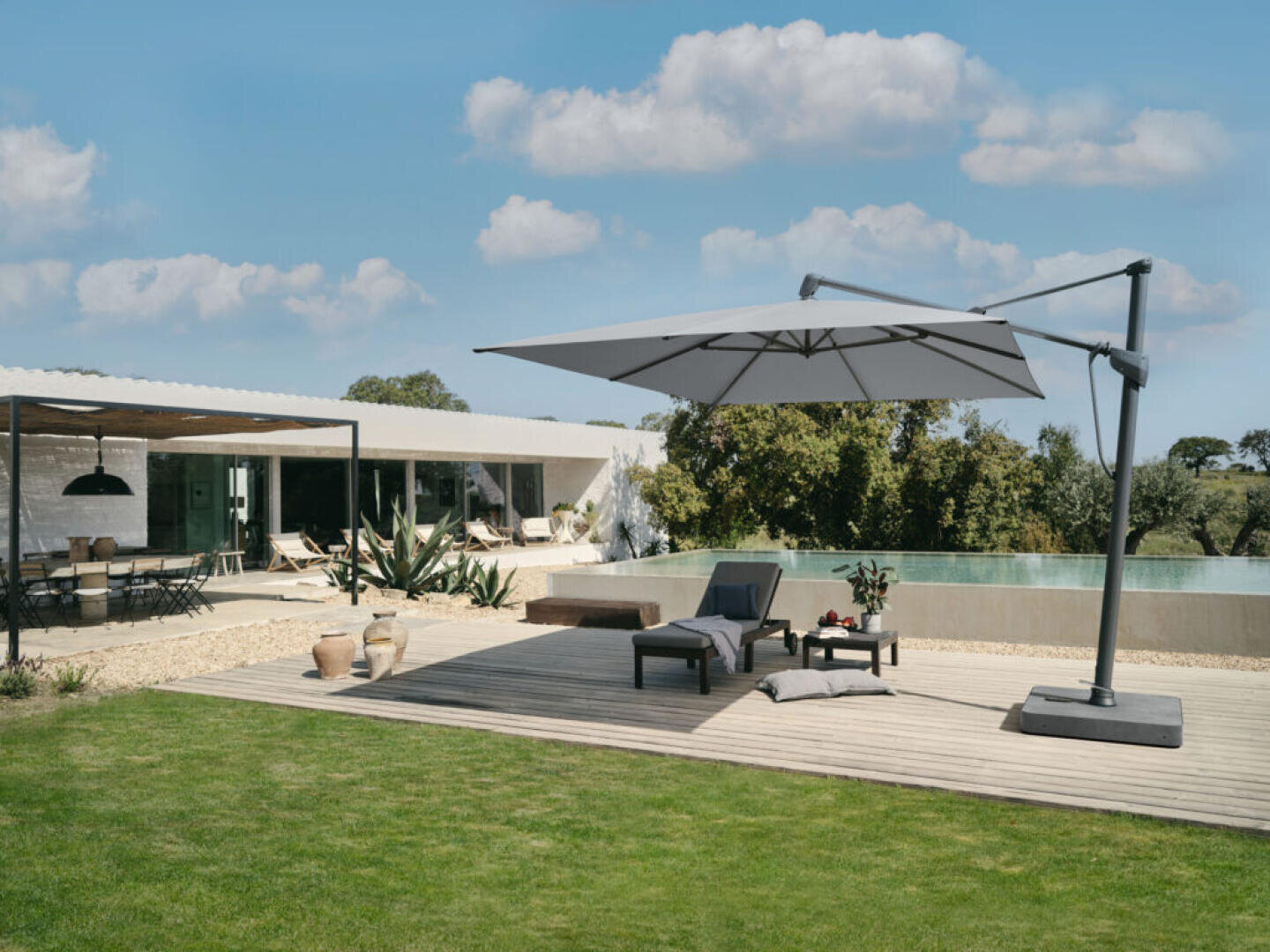 Ein moderner Garten mit einem Pool, Liegestühlen und einem großen freitragenden Sonnenschirm auf einer Holzterrasse; ein überdachter Essbereich im Freien und ein modernes Haus sind im Hintergrund unter einem teilweise bewölkten Himmel zu sehen.
