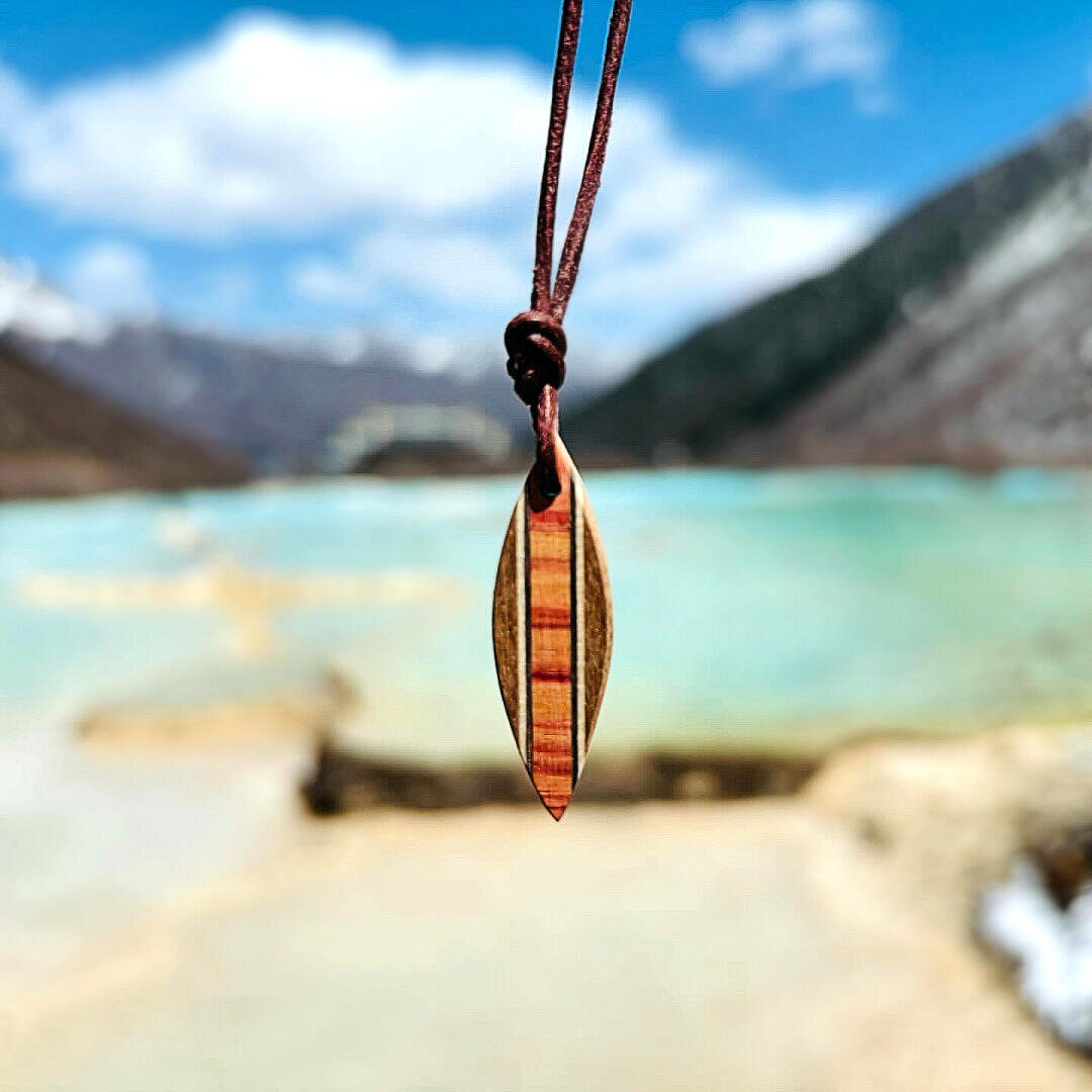 A small wooden surfboard pendant hangs from a brown cord, in the background a turquoise lake and blurred mountains under a partly cloudy sky.