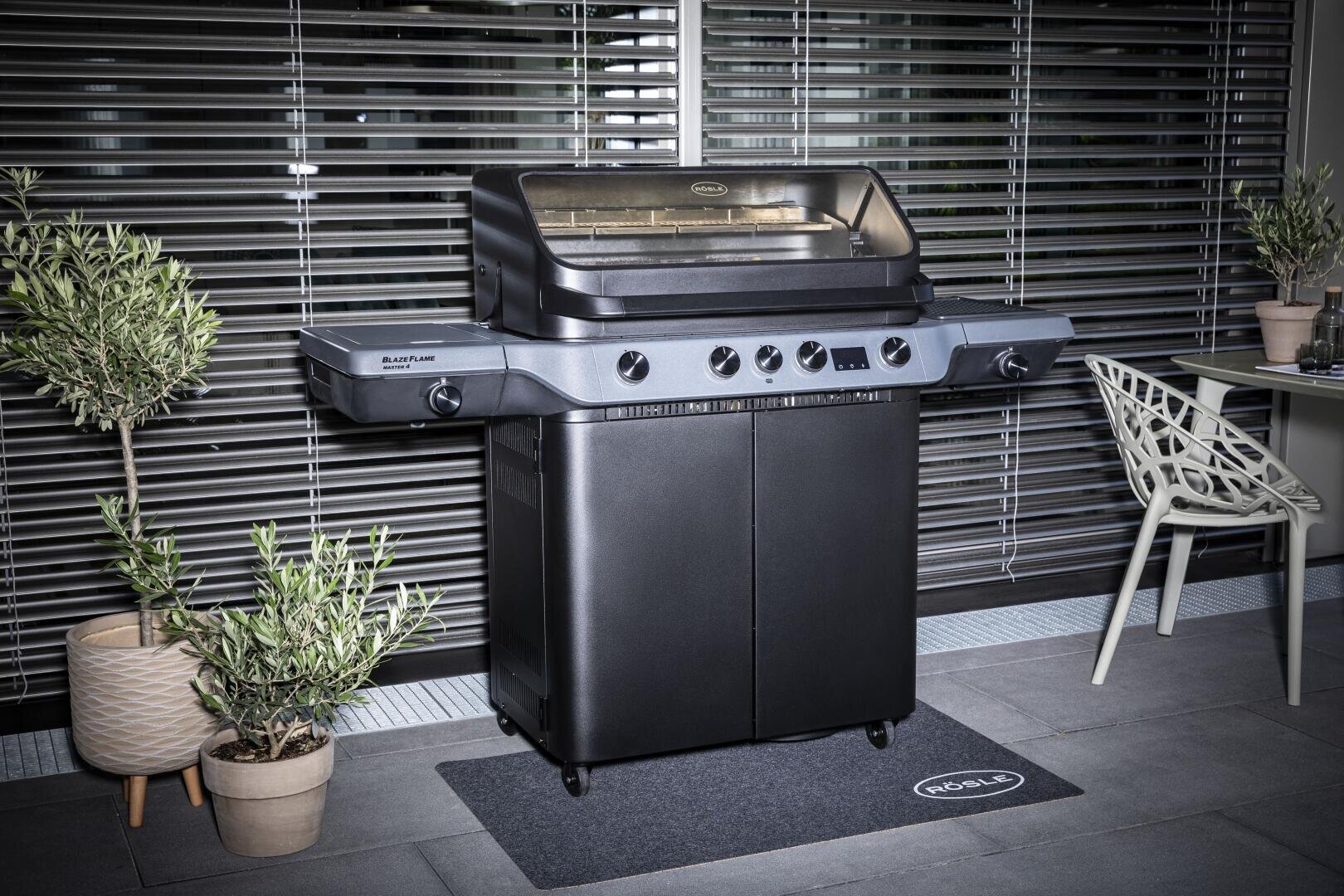 Ein schwarzer Outdoor-Gasgrill mit geschlossenem Deckel steht auf einer Matte zwischen zwei Topfpflanzen auf einer gefliesten Terrasse. In der Nähe befinden sich ein moderner Stuhl und ein kleiner Tisch, im Hintergrund sind Metalljalousien zu sehen.
