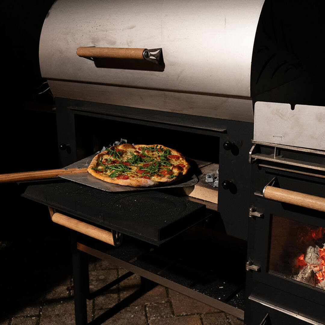 A freshly baked pizza on a wooden pizza peel being taken out of a metal outdoor pizza oven, with a fire burning in a side compartment.
