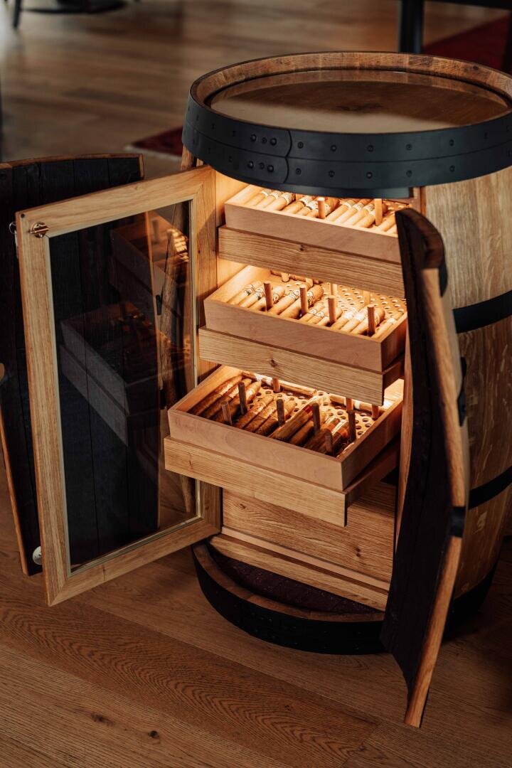Ein Humidor aus Holzfässern mit einer Glastür ist geöffnet und gibt den Blick auf mehrere beleuchtete Holzschubladen frei, die ordentlich mit Zigarren gefüllt sind und auf einem Holzboden stehen.