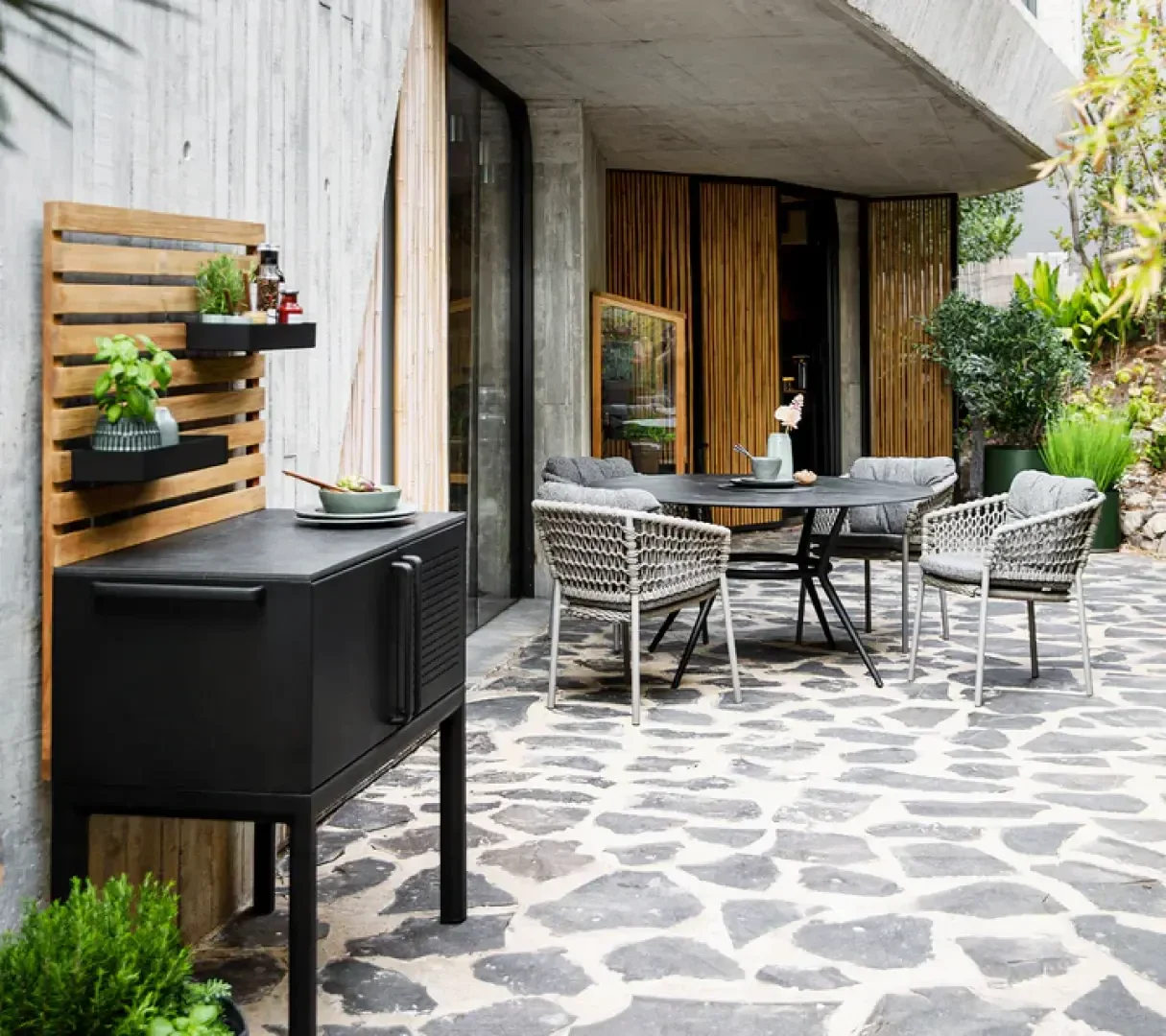 A modern patio features a round table with four woven chairs, plush cushions, and potted plants. A black outdoor grill stands nearby, with herbs and decor on a wooden slat wall. The patio floor is made of irregular stone tiles.
