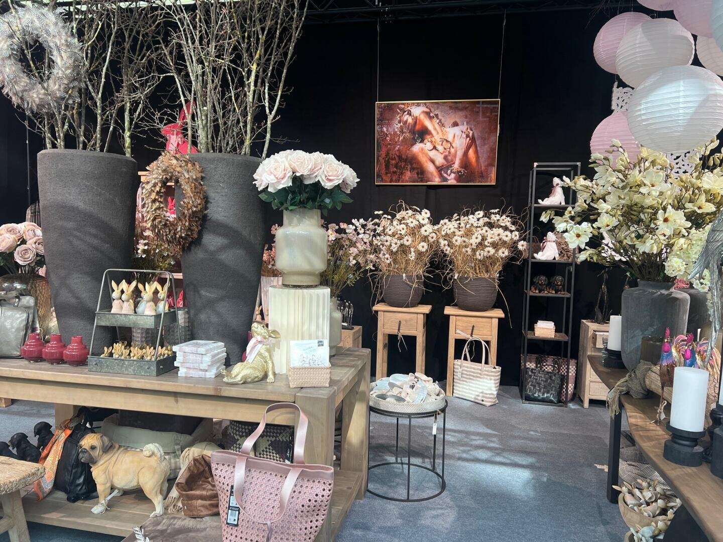 Eine Auslage mit Blumenarrangements, Vasen und Dekorationsartikeln auf Holztischen, mit Kunstblumen, Kerzen, Körben und einem Plüschhund in einer Ladeneinrichtung. Rosa und neutrale Töne dominieren die Szene.