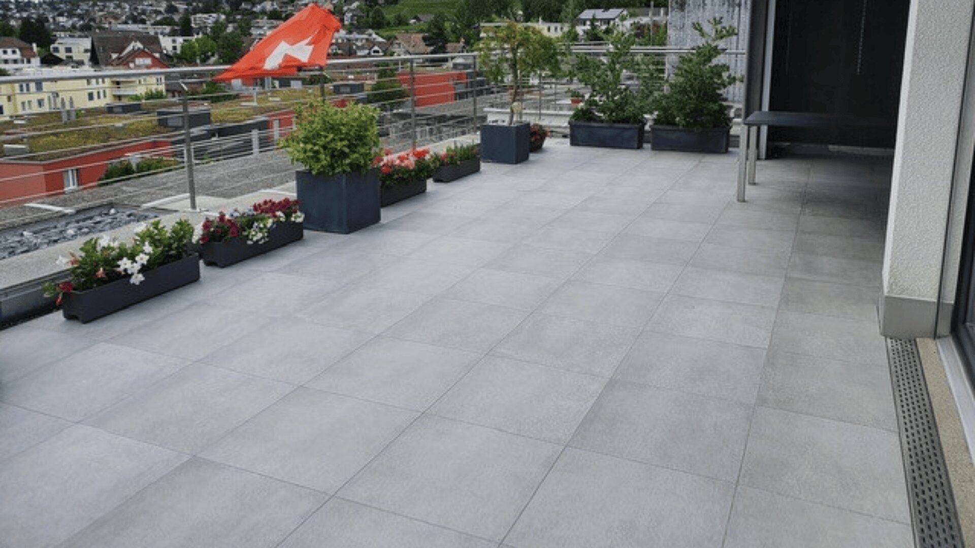 Ein geräumiger, gefliester Balkon mit Topfpflanzen und Blumen entlang des Geländers bietet einen Blick auf ein Wohngebiet mit Häusern und Grünflächen im Hintergrund.