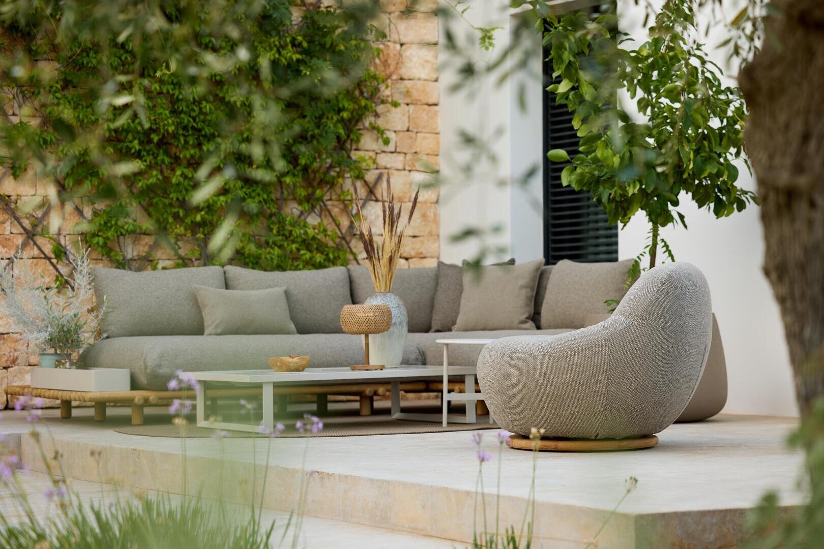 Outdoor terrace with a modern gray sectional sofa, a round armchair and low coffee tables on a stone patio surrounded by greenery and plants, next to a white brick wall.
