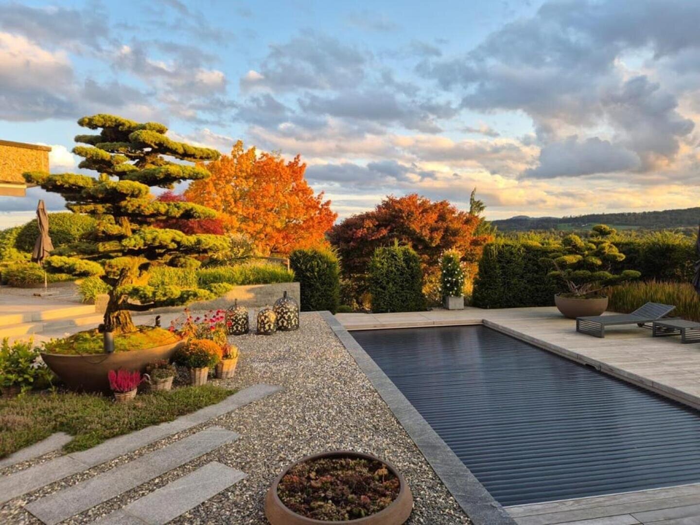 In the garden there is a covered pool, well-tended trees in large pots, colorful autumn leaves, stone steps, potted flowers and sun loungers, all under a partly cloudy sky with warm sunlight.