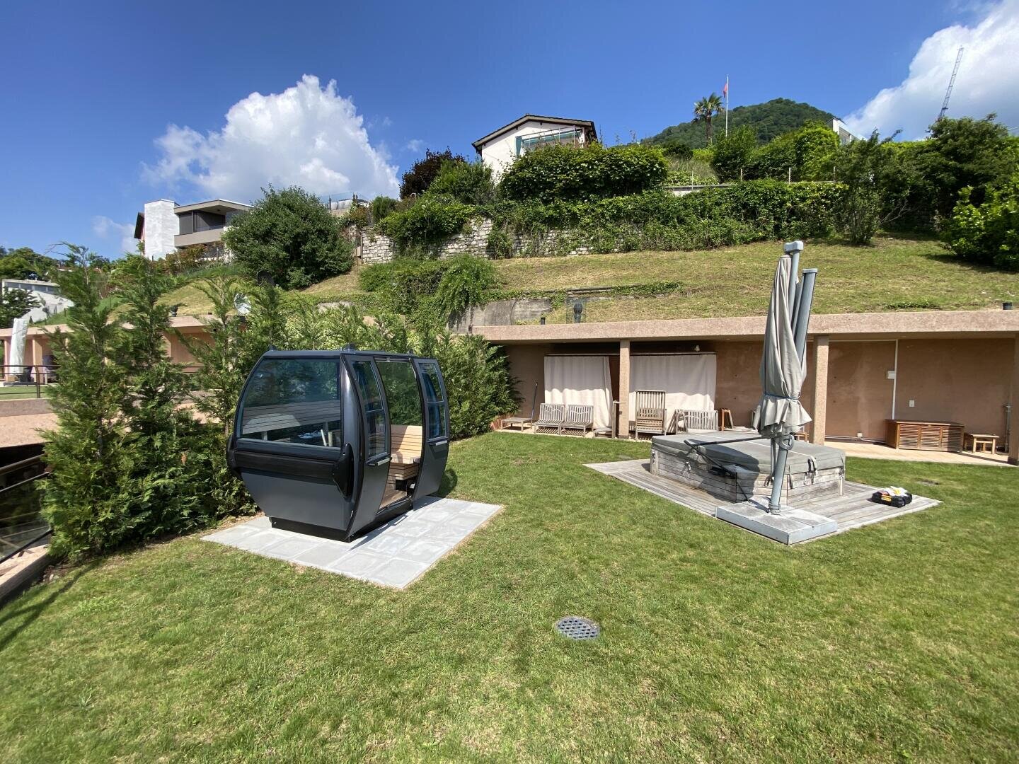 A grassy backyard with a ski gondola cabin used as a seating area, a covered outdoor patio with chairs, a closed umbrella, and hills with houses and greenery in the background under a blue sky.