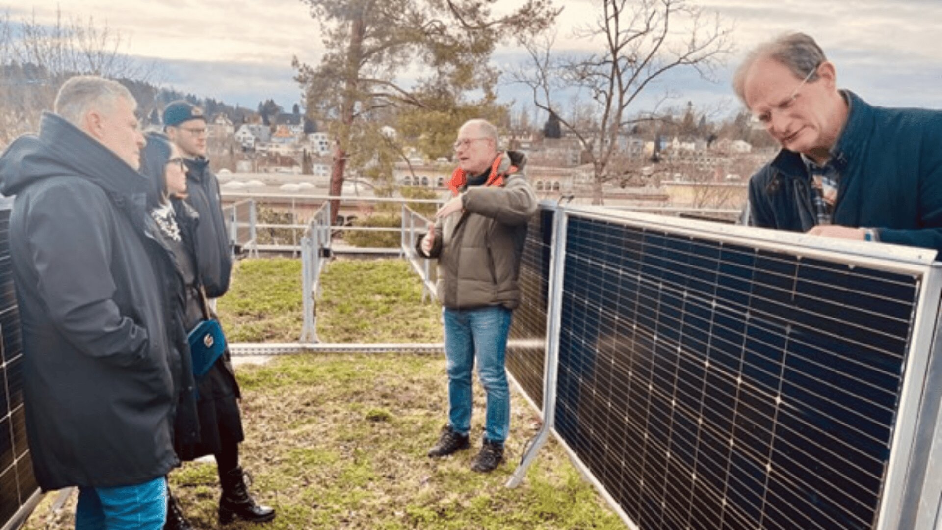 Vier Personen stehen im Freien um Solarmodule herum, während eine Person ein Modul justiert. Im Hintergrund sind Bäume und Gebäude unter einem bewölkten Himmel zu sehen.