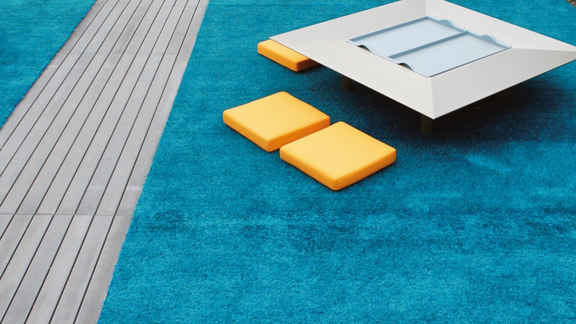 Modern outdoor space with bright blue carpet, three square yellow cushions, a low rectangular table with a glass inset, and a strip of gray wooden decking along the left side.