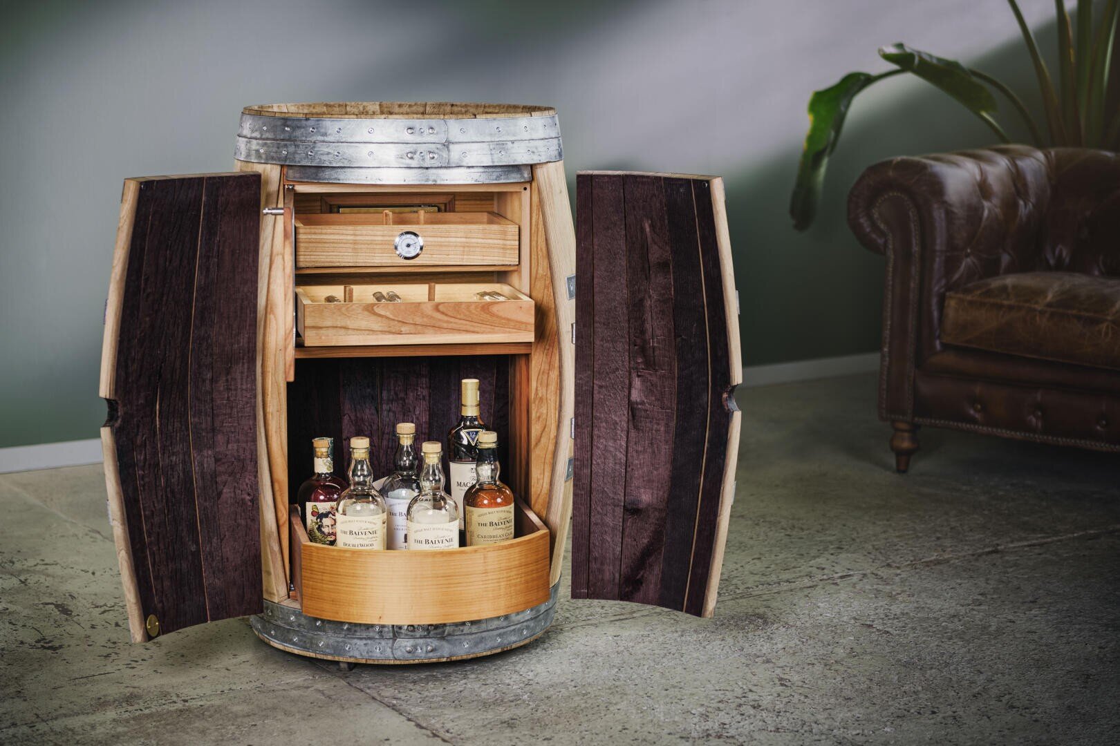 Ein Holzfass, das zu einem Schrank mit offenen Türen umfunktioniert wurde, mit Schnapsflaschen im unteren Fach und einer Schublade darüber, neben einem braunen Ledersofa und einer grünen Pflanze in einem modernen Raum.
