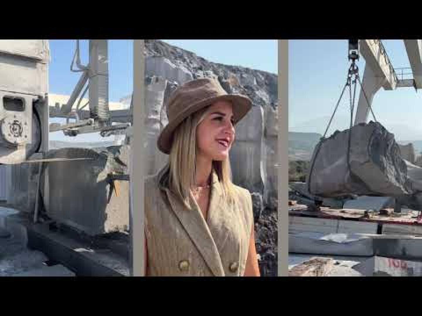 A woman wearing a tan hat and vest stands in a stone quarry, with large blocks of stone and industrial machinery visible in the background.