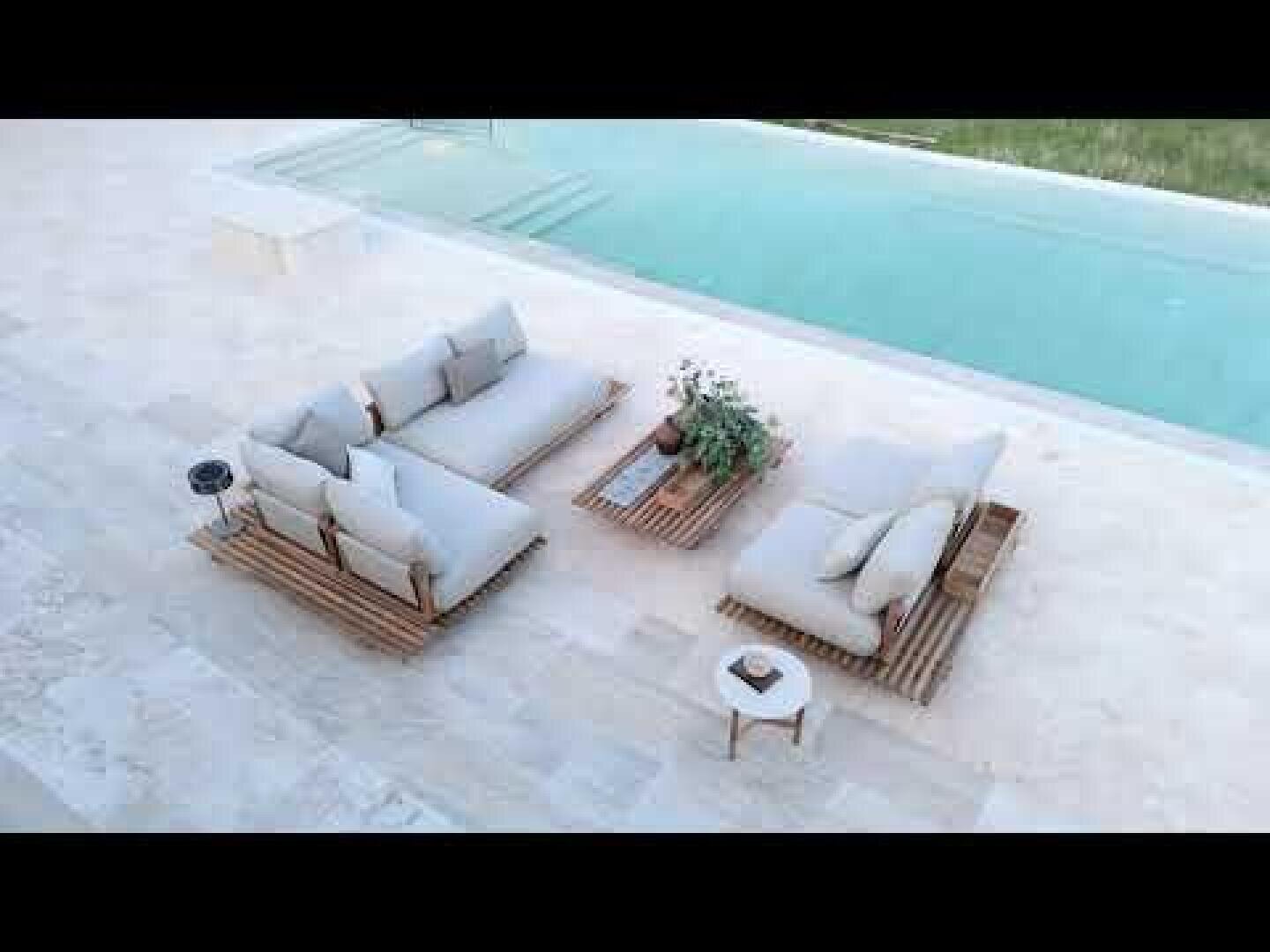 Outdoor seating area next to the pool with two upholstered sofas and a low table on a light-colored stone terrace, decorated with potted plants and a small round side table.