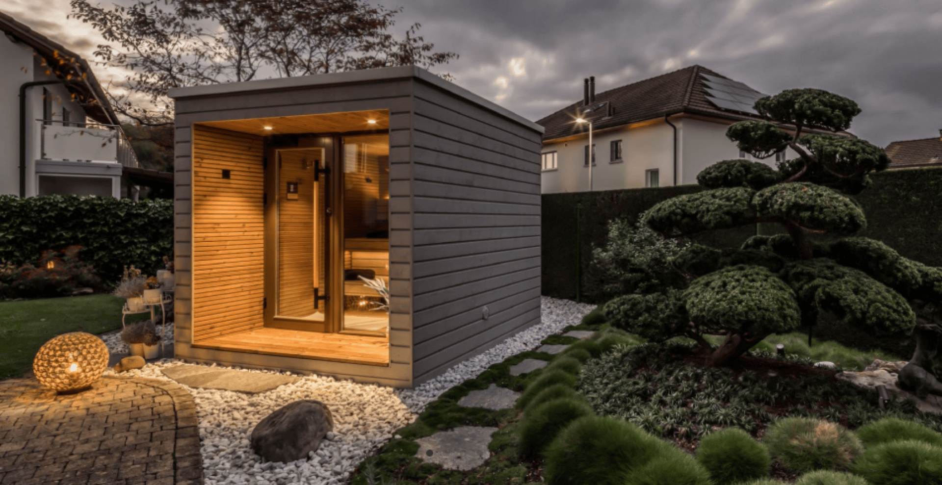 Eine moderne hölzerne Saunakabine mit warmer Innenbeleuchtung steht in einem gepflegten Garten mit ordentlich gestutzten Büschen, einem Steinweg und dekorativer Außenbeleuchtung in der Abenddämmerung.
