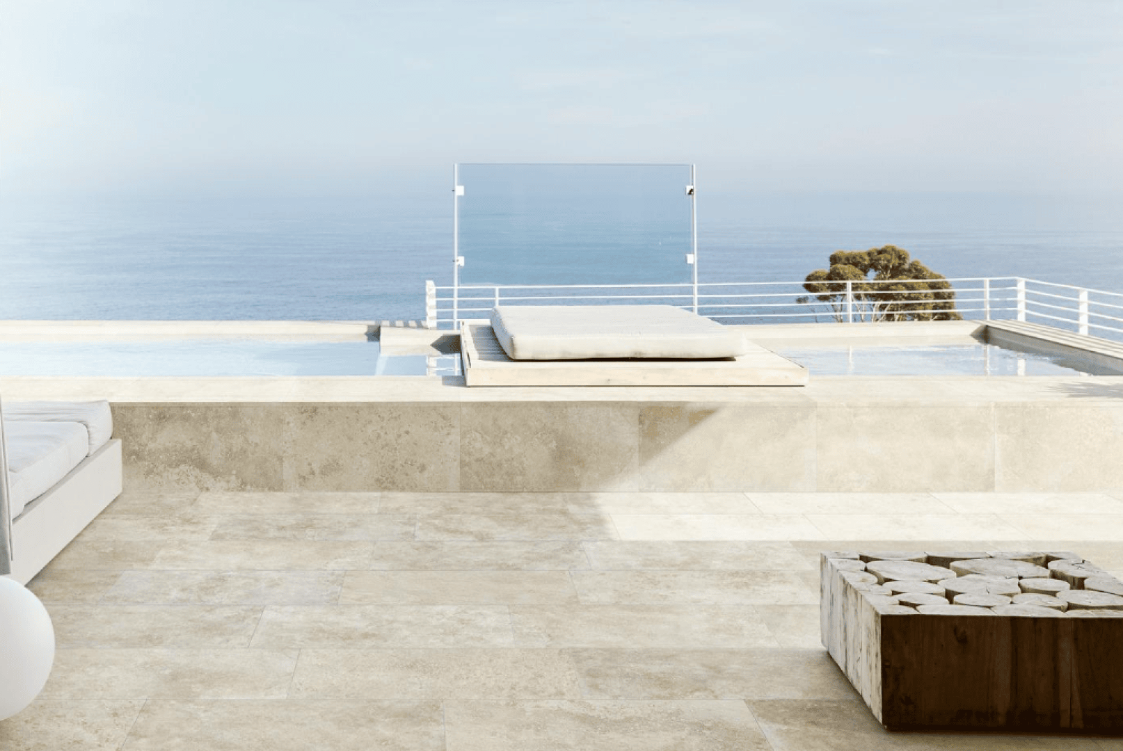 Minimalistische Terrasse mit Steinboden, einem modernen weißen Loungesessel, einem Holzblocktisch, einem Infinity-Pool und einem Glasgeländer mit Blick auf einen ruhigen Ozean unter klarem Himmel. Im Hintergrund ist ein kleiner Baum zu sehen.