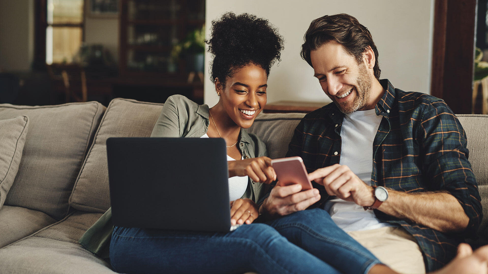 Ein lächelndes Paar sitzt auf einer Couch; die Frau hat einen Laptop auf dem Schoß und zeigt auf ein Smartphone, das der Mann in der Hand hält, beide scheinen glücklich und beschäftigt mit dem Gerät.