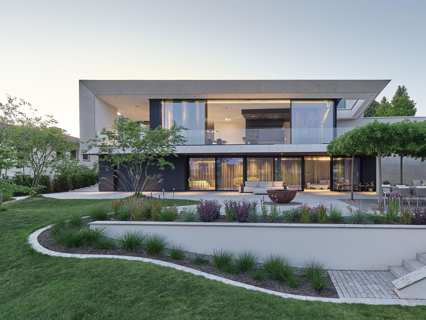 A modern two-storey house with large glass windows and a spacious terrace, surrounded by greenery and a well-tended lawn in the front.