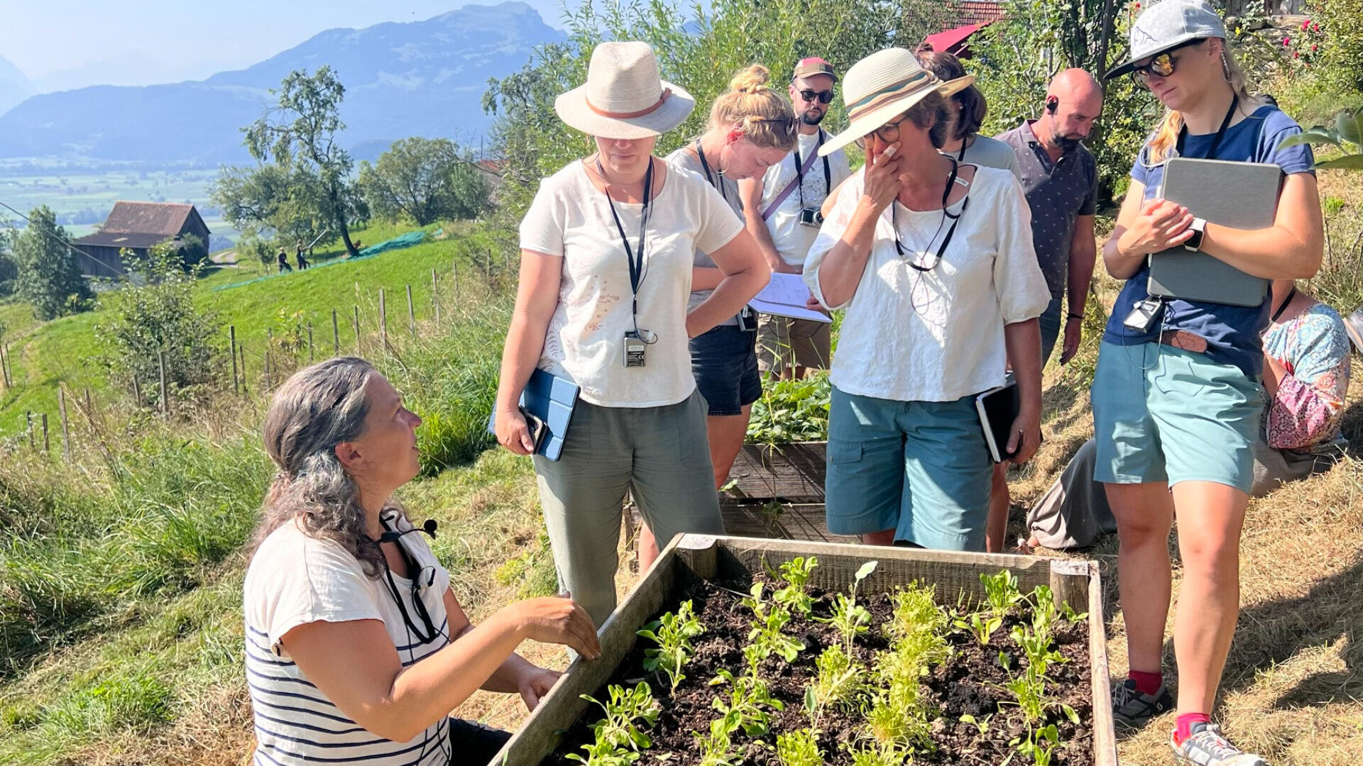Eine Gruppe von Menschen steht um ein Hochbeet im Freien und hört einer Frau mit grauen Haaren zu, die etwas über Pflanzen erklärt. Sie tragen Hüte und legere Kleidung, umgeben von Grün und Bergen im Hintergrund.
