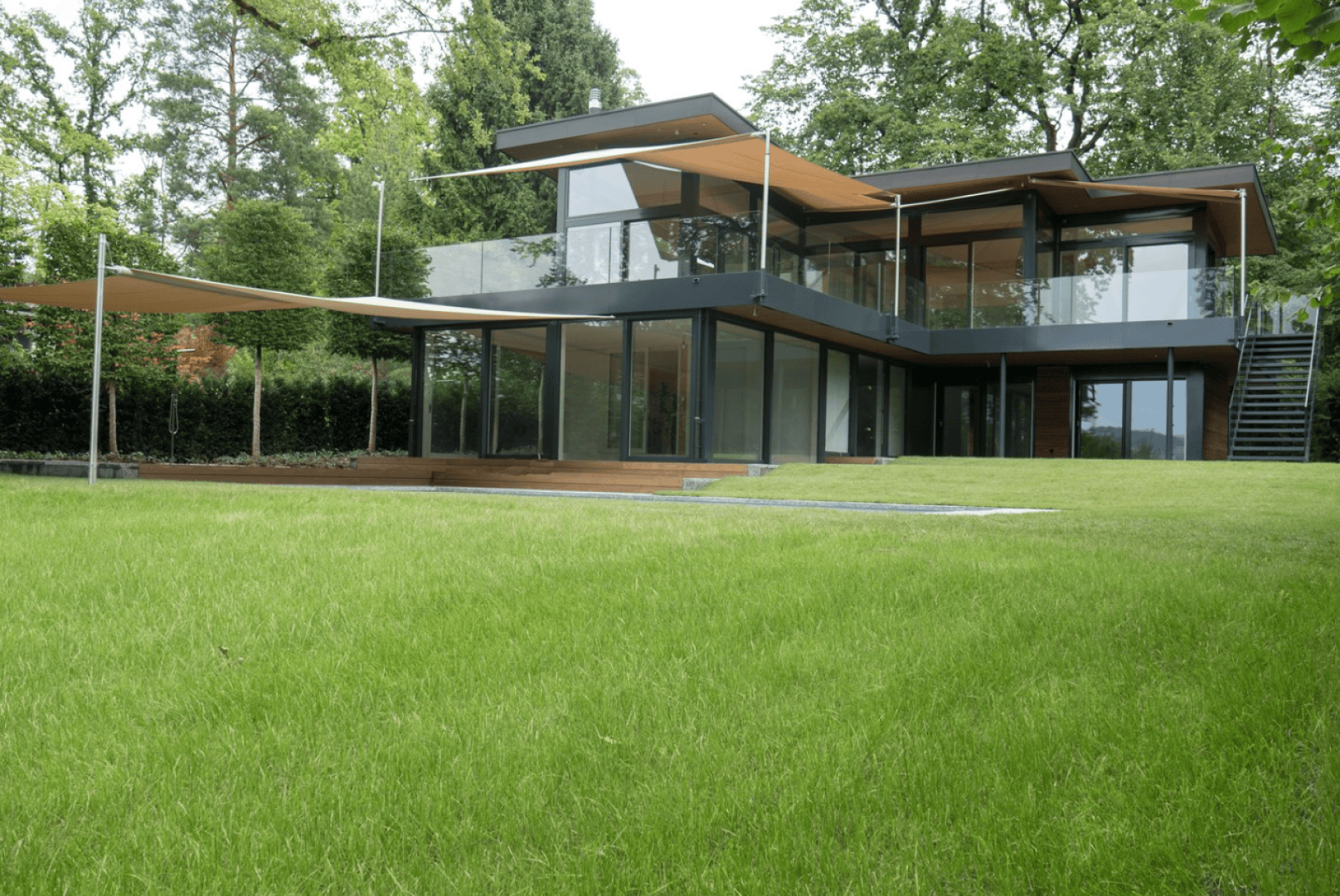 A modern two-story home with large glass windows, flat roofs and wooden accents is surrounded by lush green grass and trees. The house has outdoor terraces and several overhangs that provide shade.