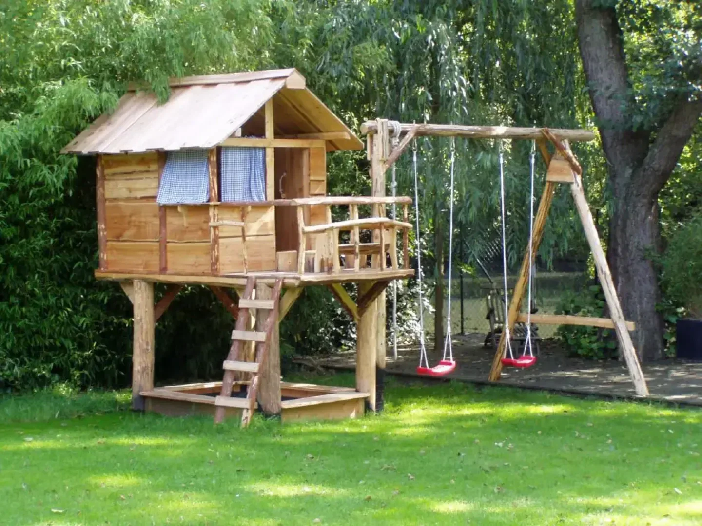 A wooden treehouse with a small porch and ladder stands next to a swing set with two red swings, surrounded by grass, trees, and greenery in a sunny backyard.