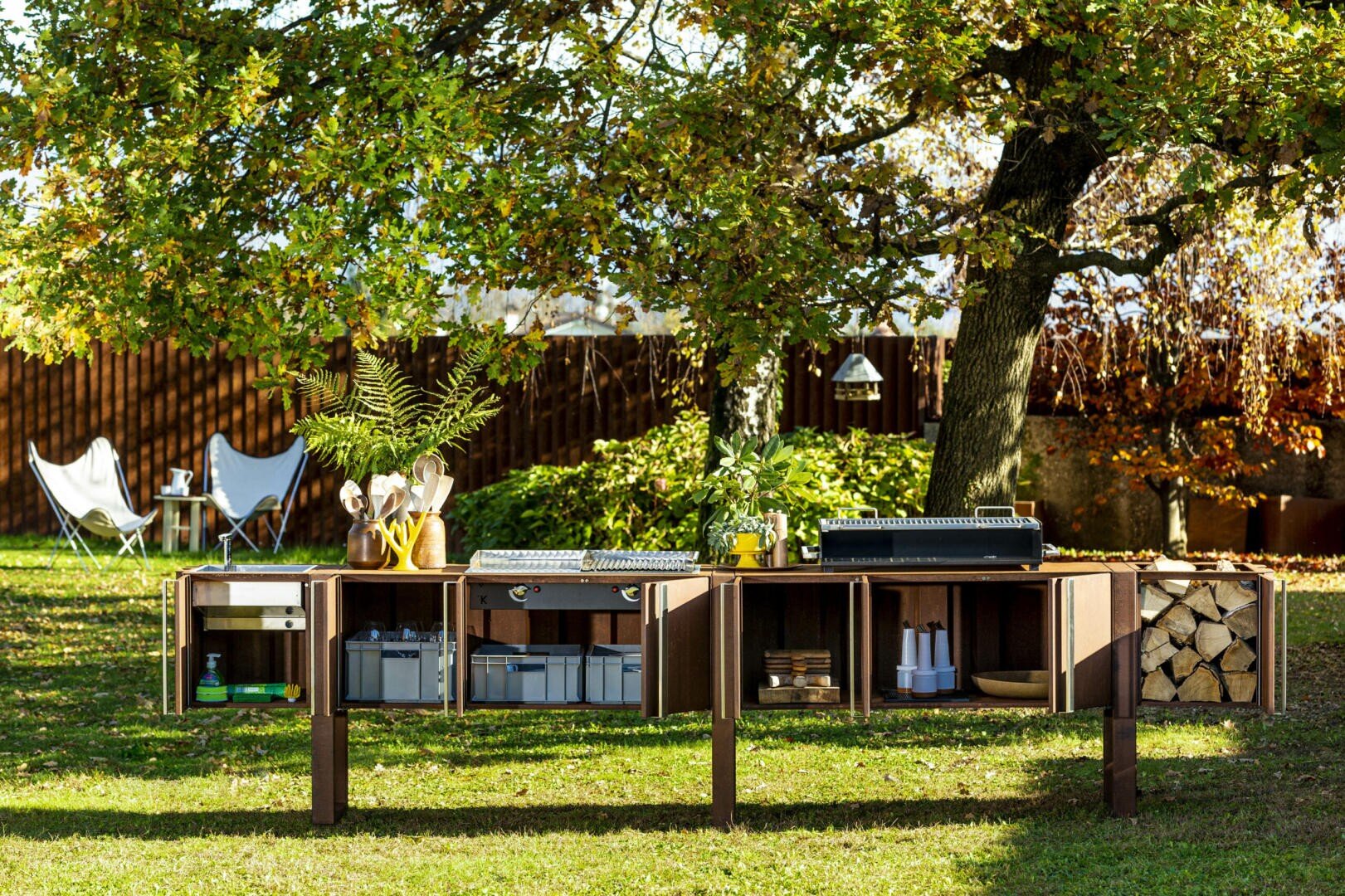 An outdoor kitchen setup under a leafy tree, featuring open shelves with kitchen supplies, potted plants, and a grill. In the background, there are chairs and a table on a grassy lawn.