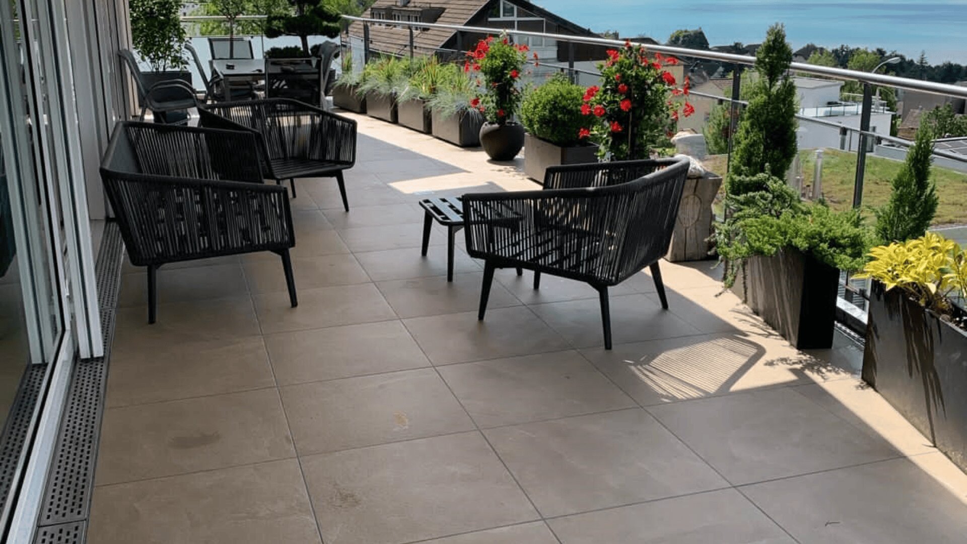 Moderner Balkon mit schwarzen geflochtenen Stühlen, kleinen Tischen und Topfpflanzen entlang des Glasgeländers. Im Hintergrund sind Häuser, Grünflächen und ein See unter einem klaren Himmel zu sehen.