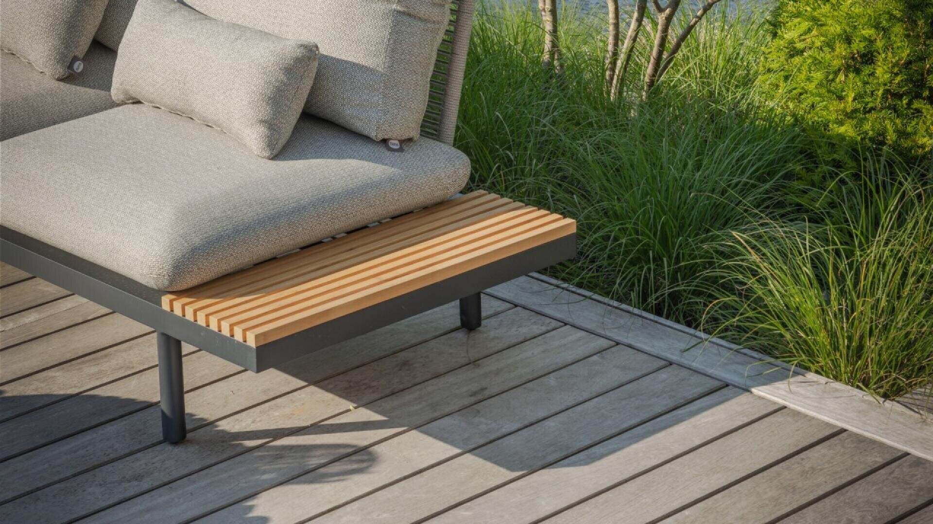 A modern outdoor lounge chair with light gray cushions and a wooden slatted side table sits on a light wood deck, surrounded by green grass and plants.