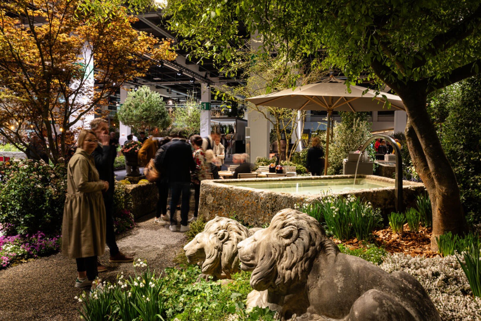 In einem üppigen Innengarten stehen zwei steinerne Löwenstatuen neben farbenfrohen Pflanzen, einem kleinen Springbrunnen und Menschen, die bei gedämpfter Beleuchtung und unter einem großen Sonnenschirm die Auslagen bewundern.