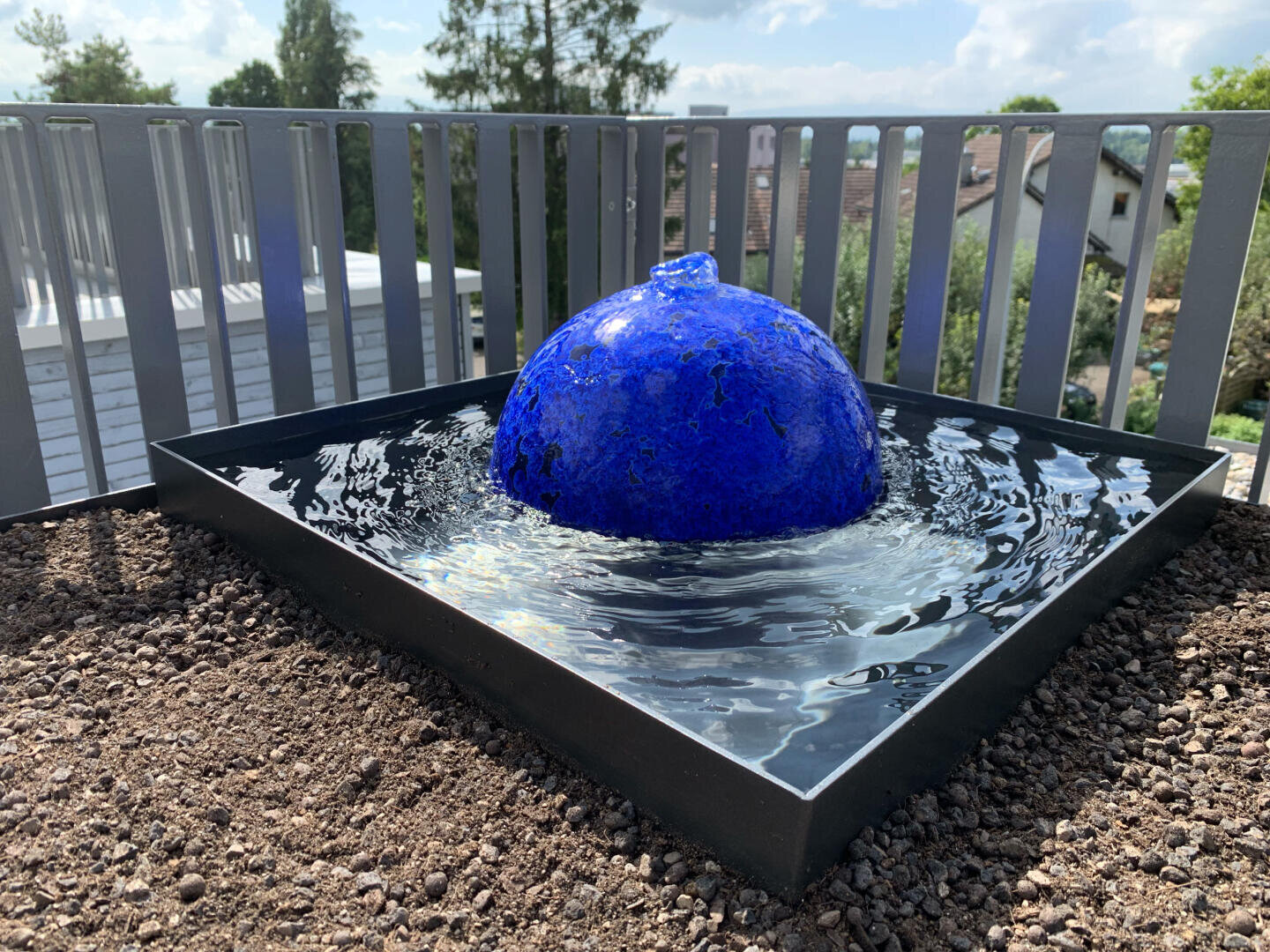 Ein leuchtend blauer, kugelförmiger Springbrunnen sitzt in einem quadratischen, mit Wasser gefüllten schwarzen Becken, umgeben von Kies, auf einer Dachterrasse mit Metallgeländer und Bäumen im Hintergrund.