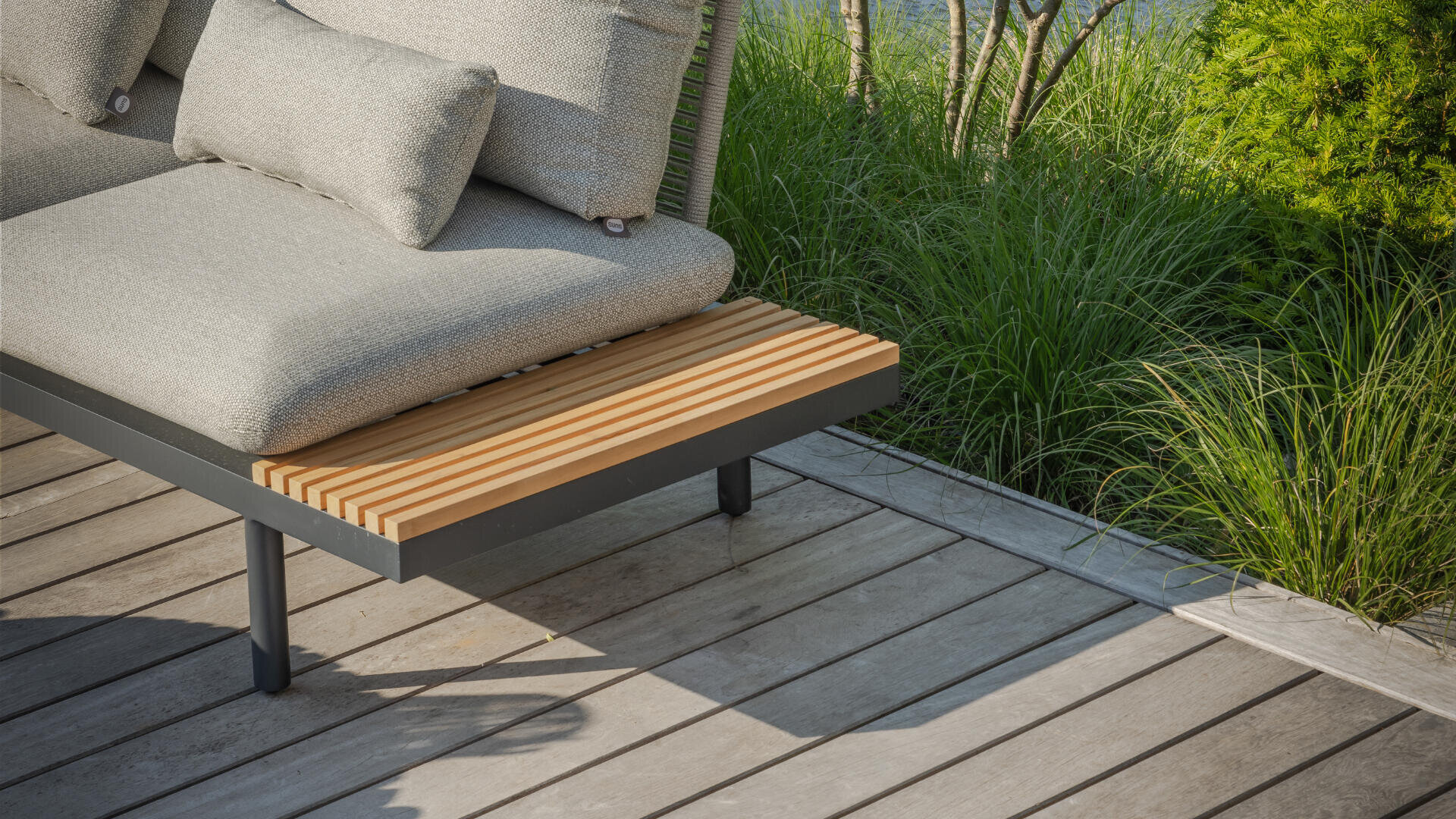 Moderne Lounge-Sitzmöbel mit hellgrauen Kissen und einem Beistelltisch aus Holzlatten auf einer Holzterrasse, umgeben von üppig grünem Gras und Sträuchern.