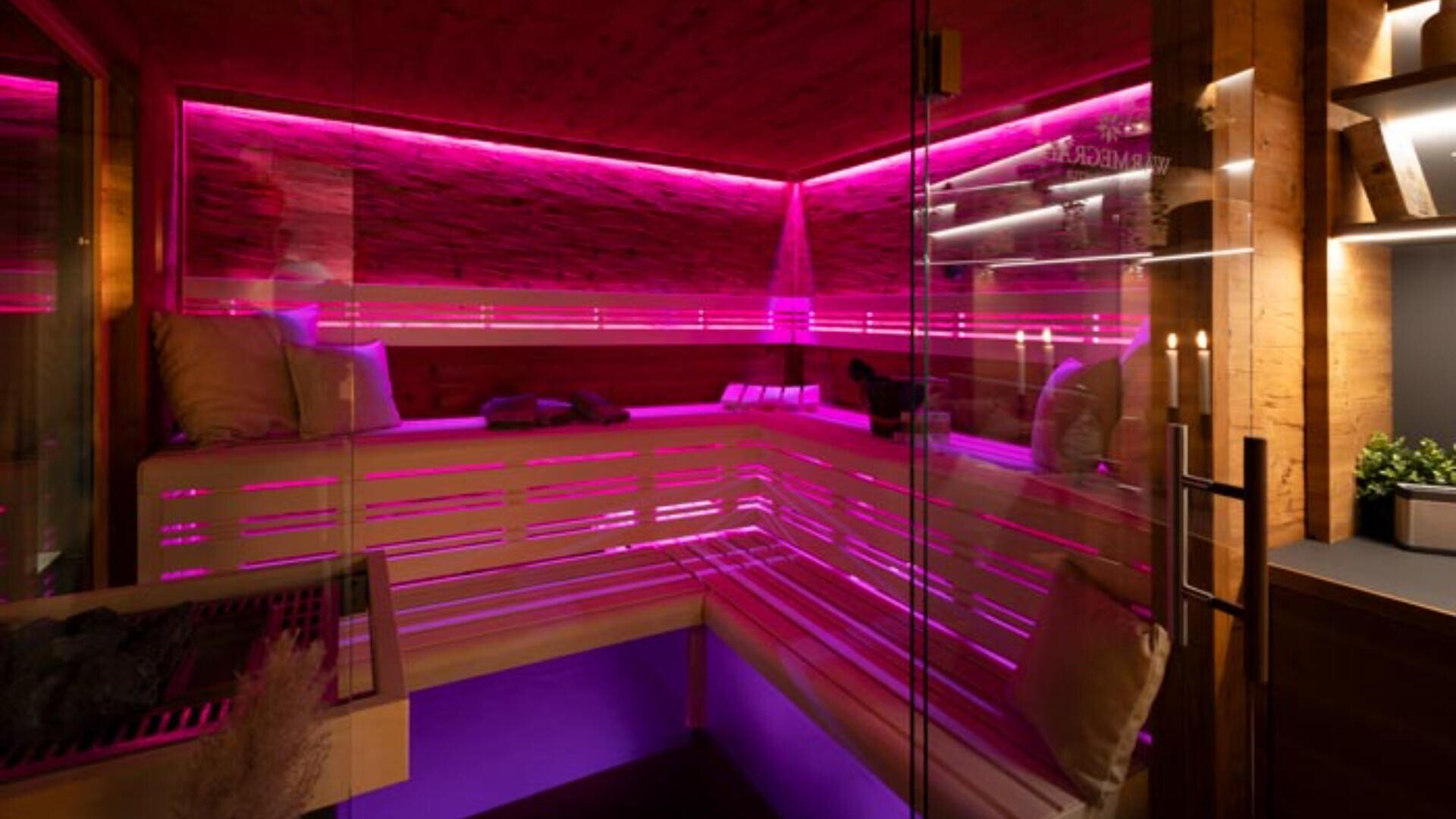 Eine moderne Sauna mit Holzbänken, Glastüren und leuchtend pinkfarbener LED-Beleuchtung, die eine entspannende und moderne Atmosphäre schafft. Die Bänke sind mit Kissen ausgestattet, die für Komfort sorgen.