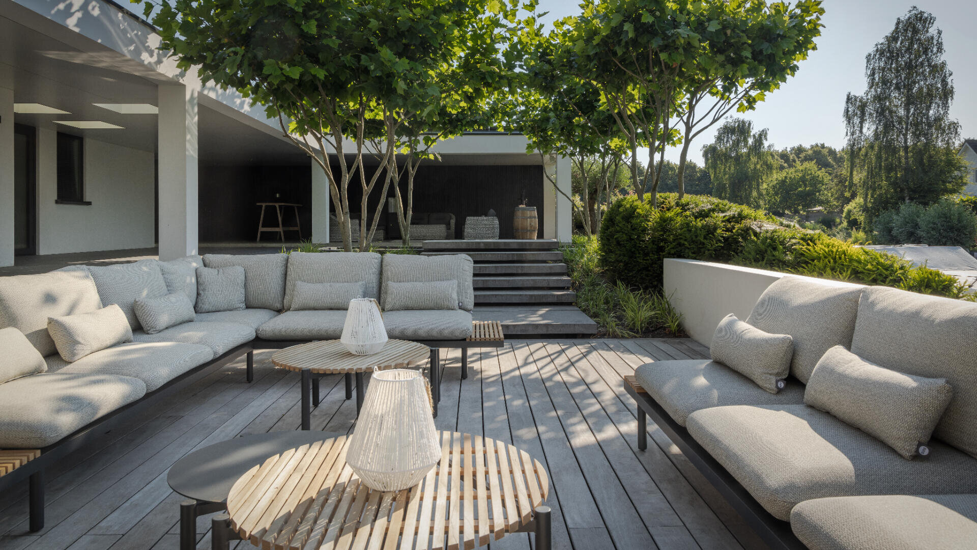 Eine moderne Außenterrasse mit hellgrauen Sofas, runden Holztischen und dekorativen Laternen, umgeben von üppigem Grün und Bäumen, mit einem überdachten Sitzbereich im Hintergrund.