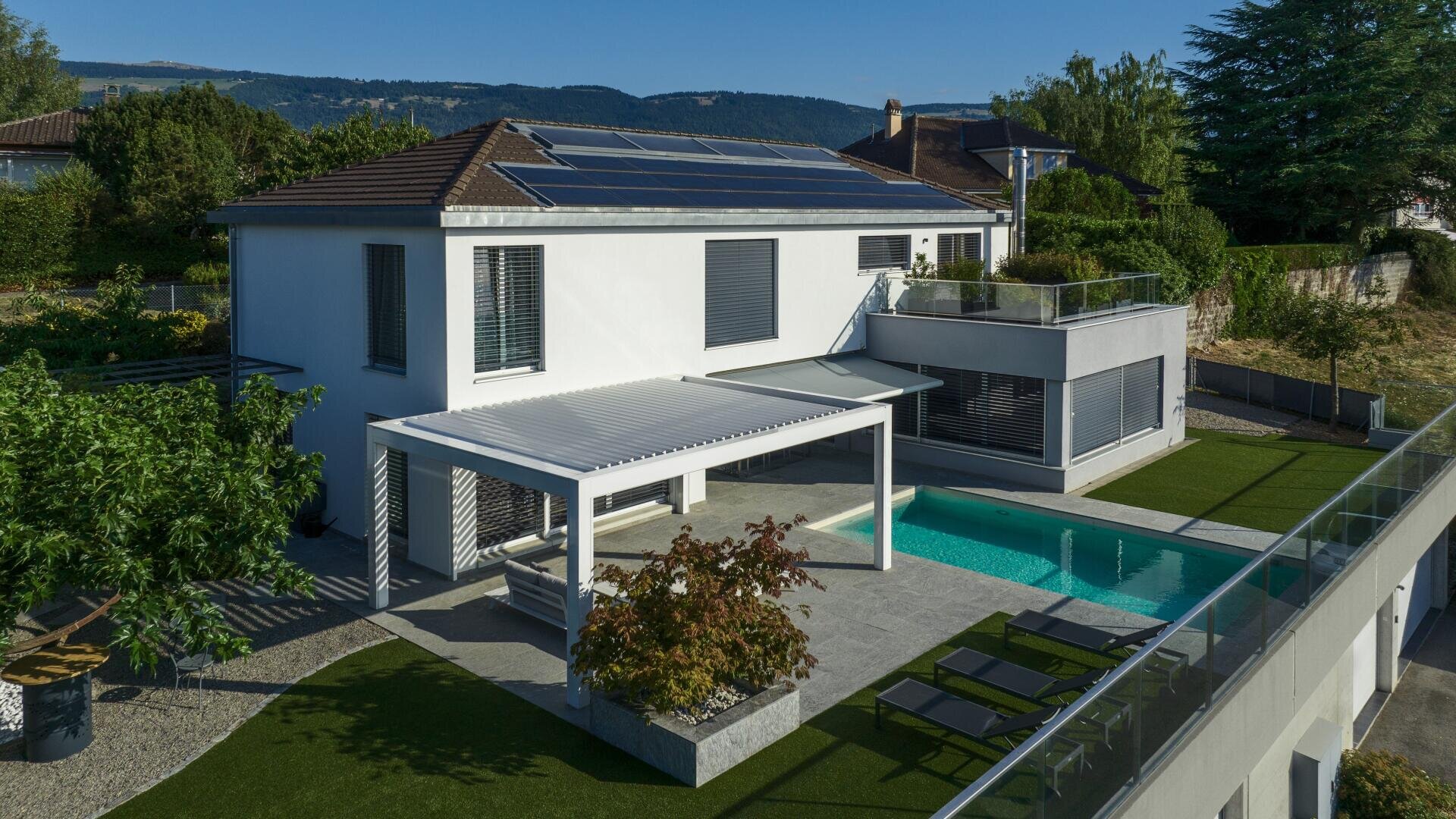 Modern two-story house with solar panels on the roof, a patio with a pergola, an outdoor swimming pool, sun loungers, and a garden, set in a green, hilly neighborhood on a sunny day.