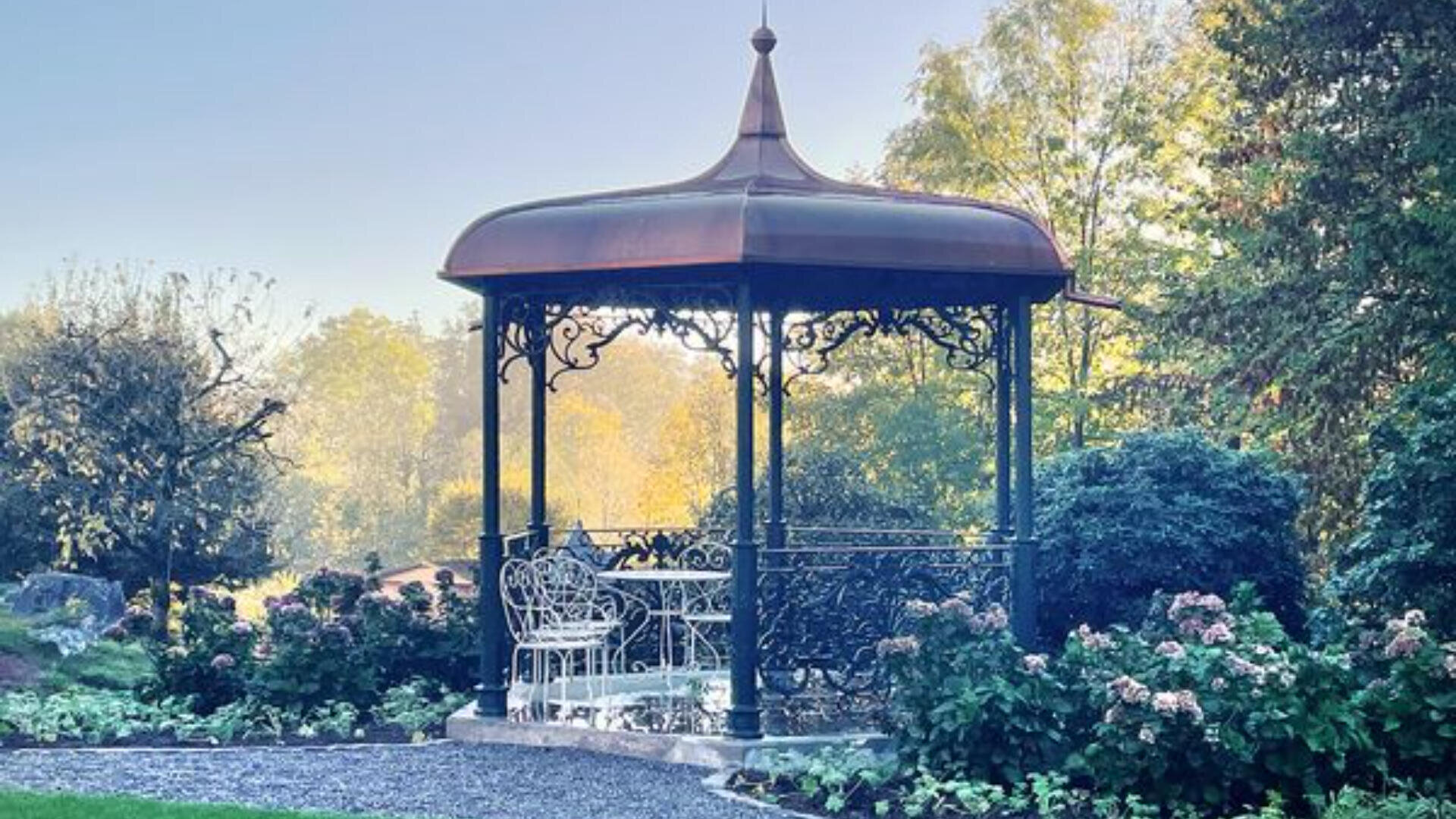Ein dekorativer Pavillon mit einem Metalldach und kunstvollen Details steht in einem Garten, umgeben von Büschen und Blumen. Im Inneren befinden sich ein weißer Metalltisch und Stühle. Das Sonnenlicht fällt durch die Bäume im Hintergrund.