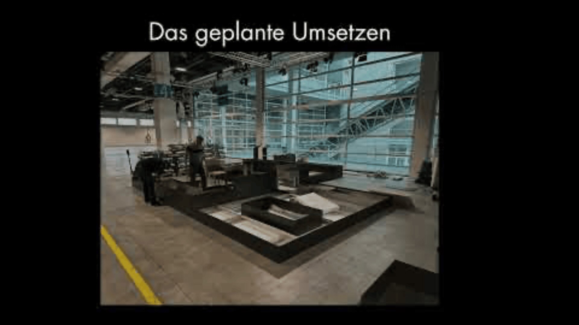 Ein moderner Innenraum mit großen Fenstern und Metallträgern, der eine teilweise montierte dunkel gefärbte Plattformstruktur aufweist. Der deutsche Text "Das geplante Umsetzen" erscheint im oberen Bereich.