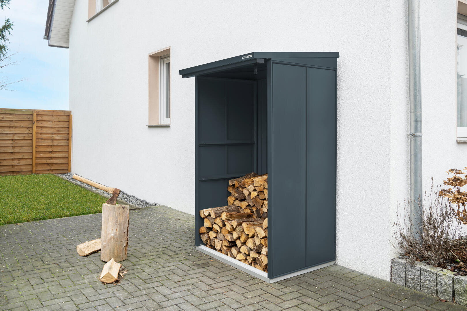 An einer weißen Außenwand steht ein kleiner metallener Holzschuppen, in dem sich gestapeltes Brennholz befindet. Einige gespaltene Holzscheite und ein Hackklotz liegen auf der grauen Steinterrasse daneben. Im Hintergrund sind Gras und ein Holzzaun zu sehen.