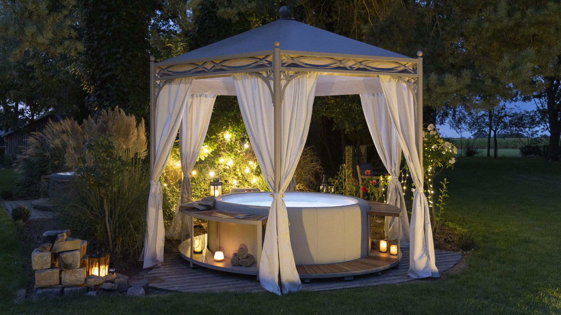 A round whirlpool under a pavilion with white curtains, surrounded by glowing lanterns and fairy lights, in the middle of a lush garden at dusk.