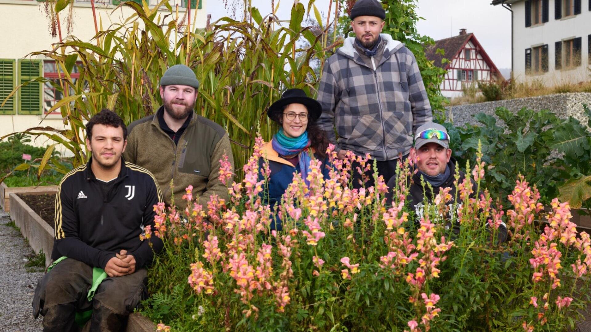 Five people pose together in a garden filled with pink and yellow flowers, with tall plants and traditional houses in the background. They are dressed warmly, and appear happy and relaxed.