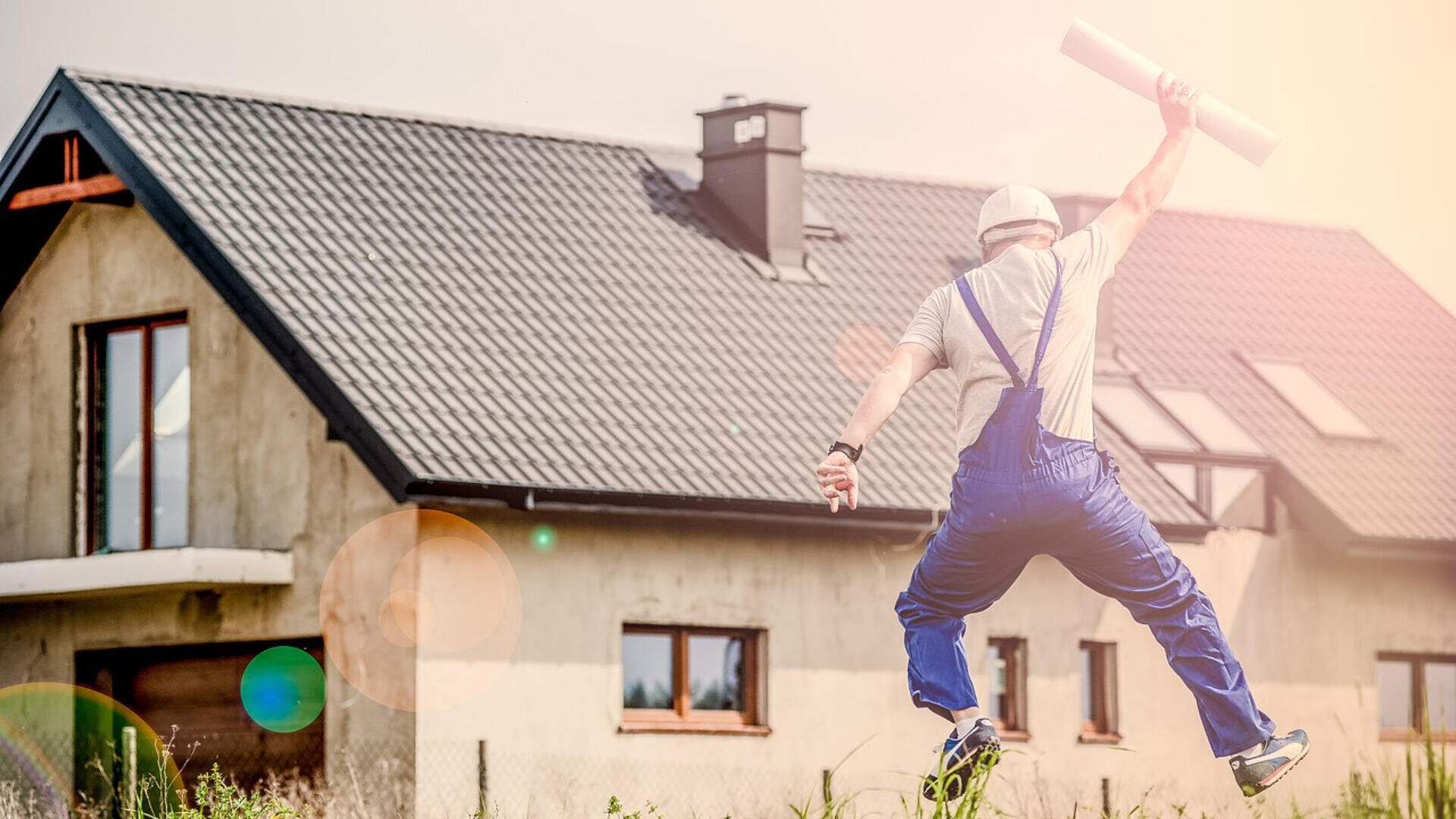 Ein Bauarbeiter in blauer Latzhose und weißem Helm springt freudig mit aufgerollten Plänen vor einem neu gebauten Haus mit dunklem Dach herum.