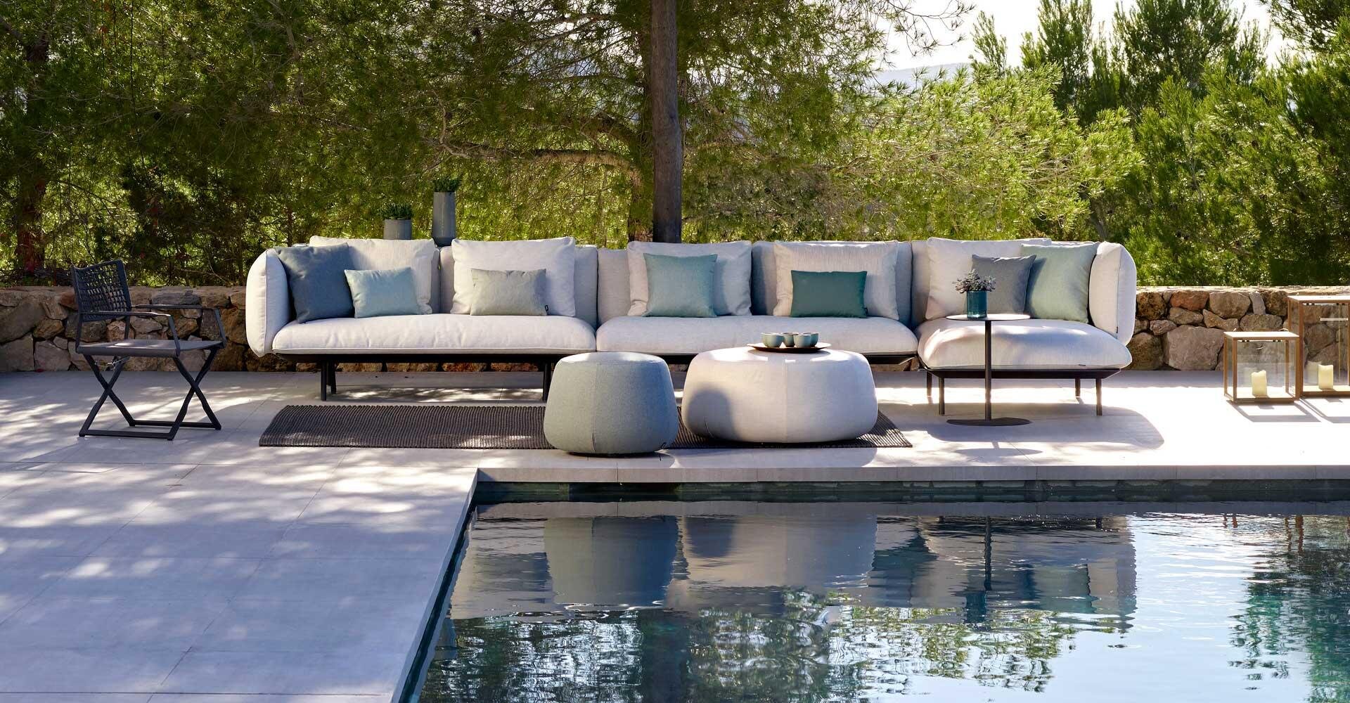 Outdoor terrace with a large white sectional sofa and blue cushions next to a swimming pool. Two round stools, a small side table, a folding chair and green trees create a relaxed, modern atmosphere.