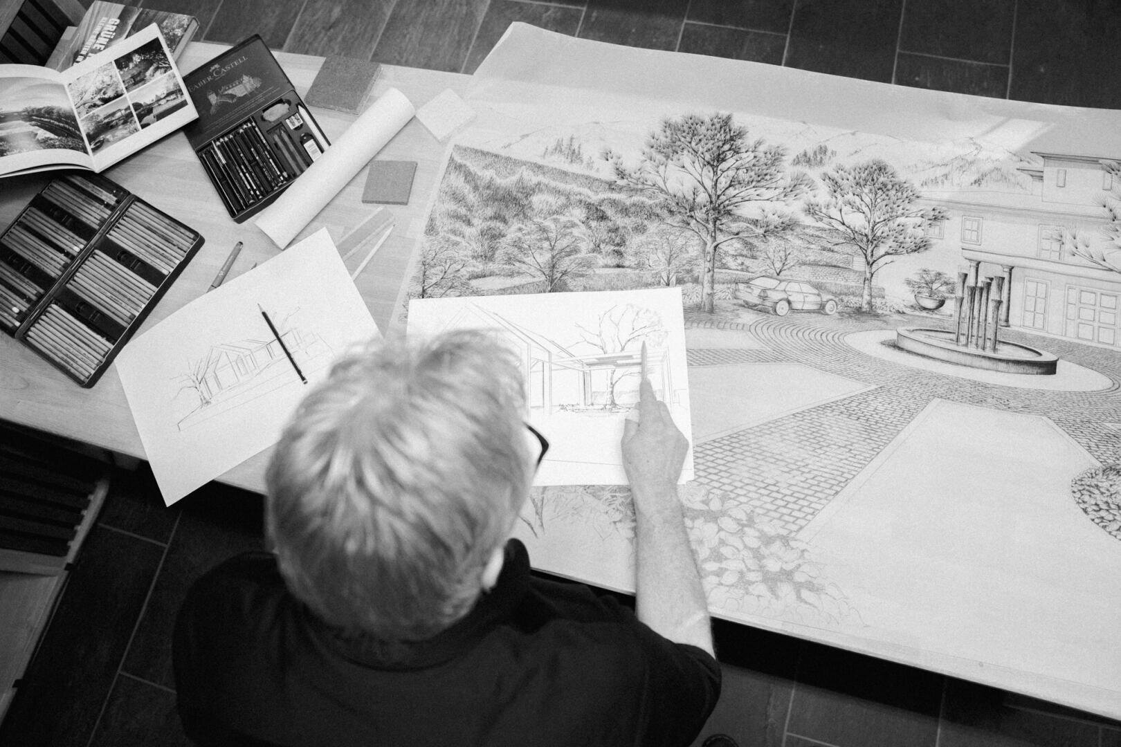 A person with light-colored hair is working on an architectural drawing and holding a sketch over a large, detailed landscape. Painting utensils and other sketches are scattered on the desk next to him.