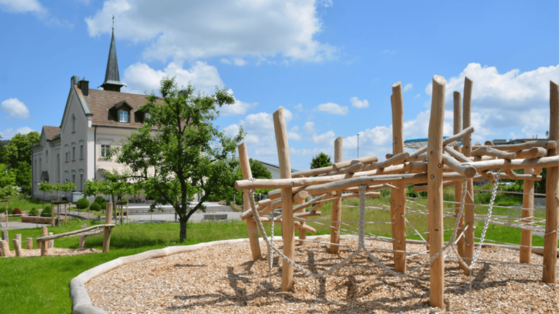 Ein hölzernes Spielplatz-Klettergerüst steht auf Mulch in einem grasbewachsenen Park, mit einem grossen weissen Gebäude und einem Kirchturm im Hintergrund unter einem blauen Himmel mit vereinzelten Wolken.