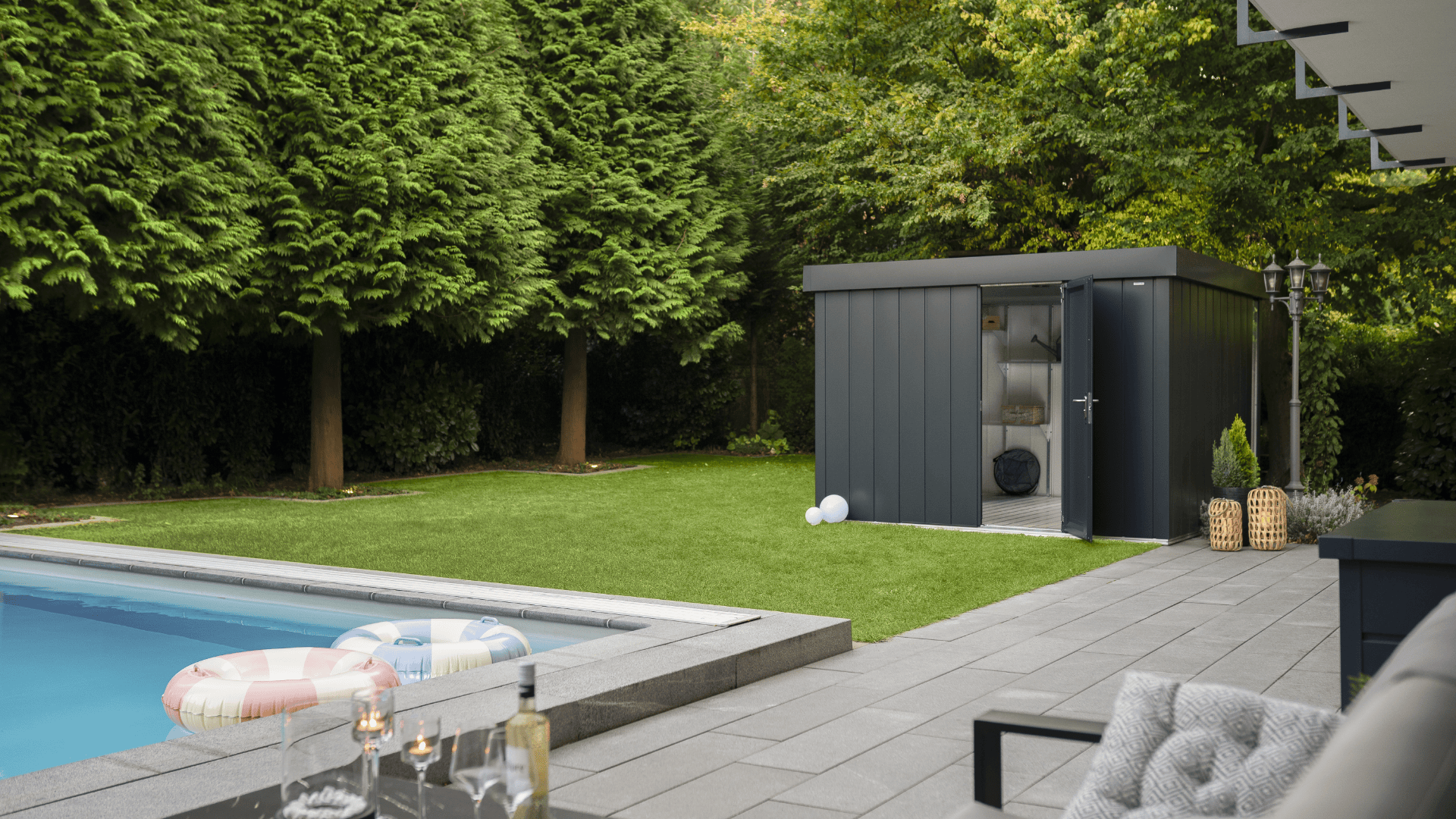 Ein moderner Hinterhof mit Rasen, einem rechteckigen Pool mit Schwimmkörpern und einem schlichten grauen Schuppen. Bäume säumen den Garten, und auf der Terrasse stehen Gartenmöbel und ein kleiner Tisch mit Getränken.