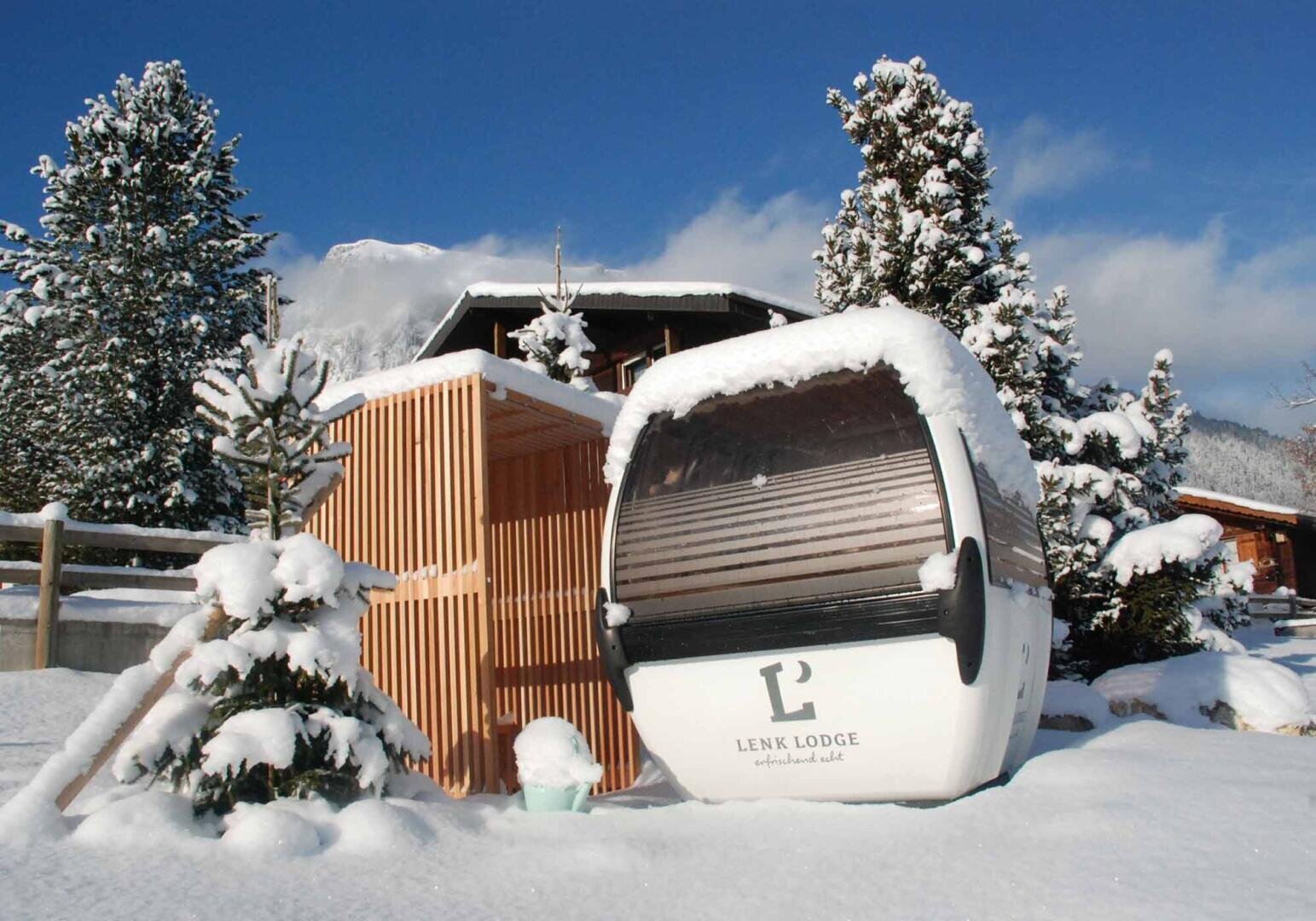 A snowy scene with a small gondola cabin marked Lenk Lodge beside a wooden structure, surrounded by snow-covered trees and mountains in the background.
