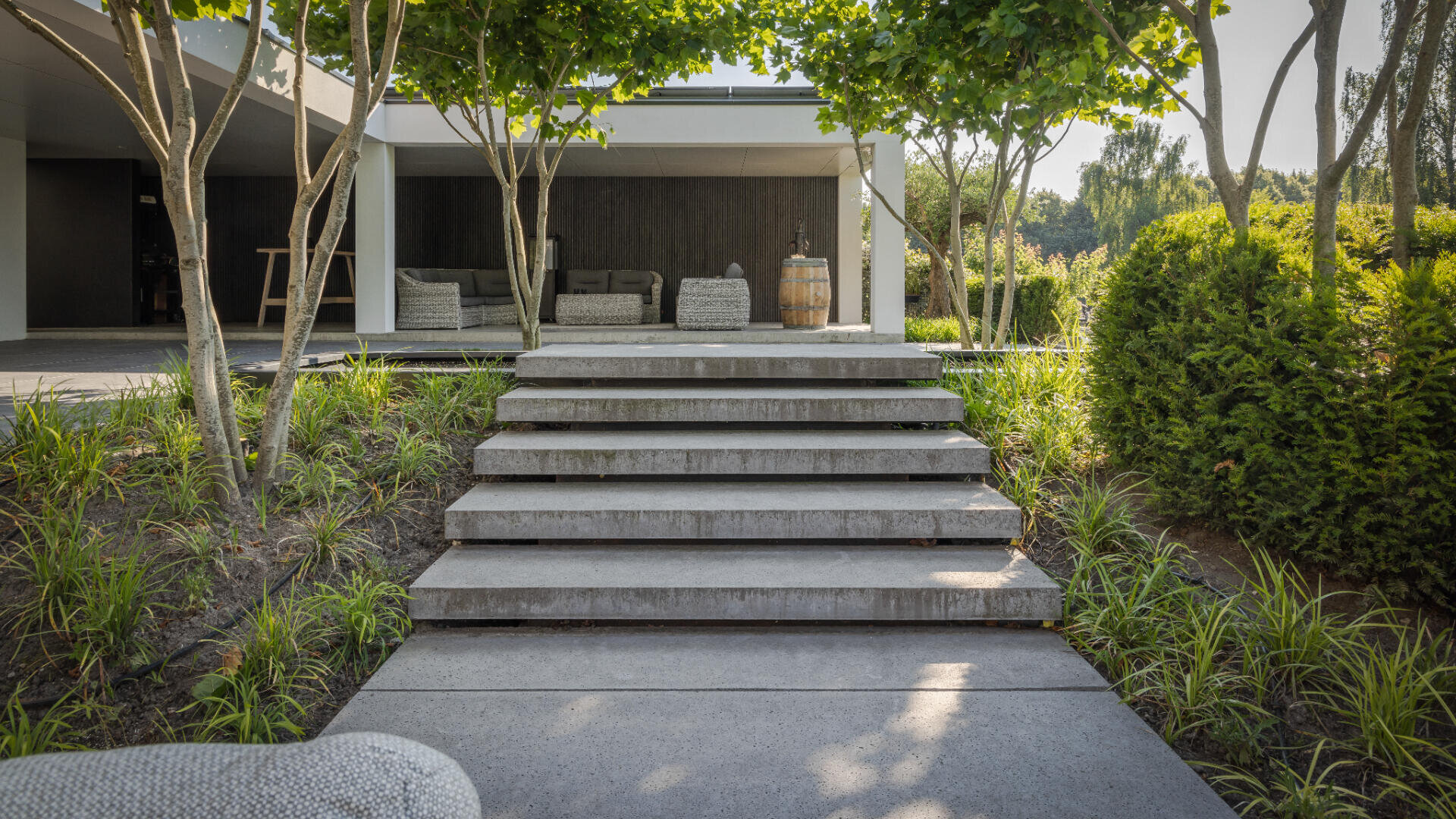 Betontreppen führen durch einen angelegten Garten mit Bäumen und Sträuchern zu einem modernen Freisitz mit Sofas unter einer überdachten Veranda.