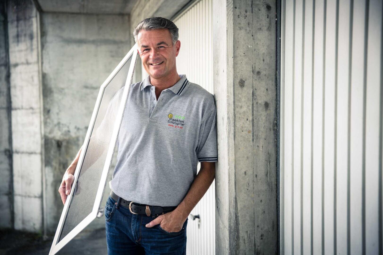 Ein lächelnder Mann in einem grauen Poloshirt steht an einer Betonwand und hält einen rechteckigen Bildschirm oder Rahmen. Er hat eine Hand in der Tasche und lehnt sich lässig gegen die Wand.