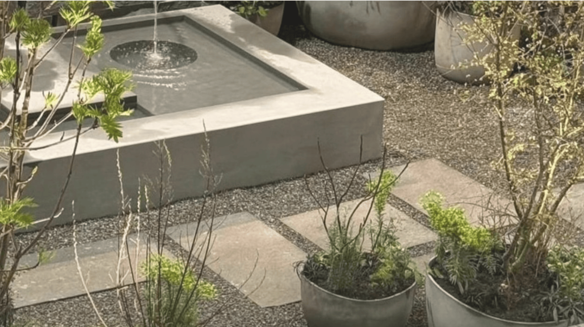 Ein moderner quadratischer Wasserbrunnen mit zentralem Auslauf ist von Kies, Pflastersteinen und grünen Topfpflanzen in einem Garten umgeben.