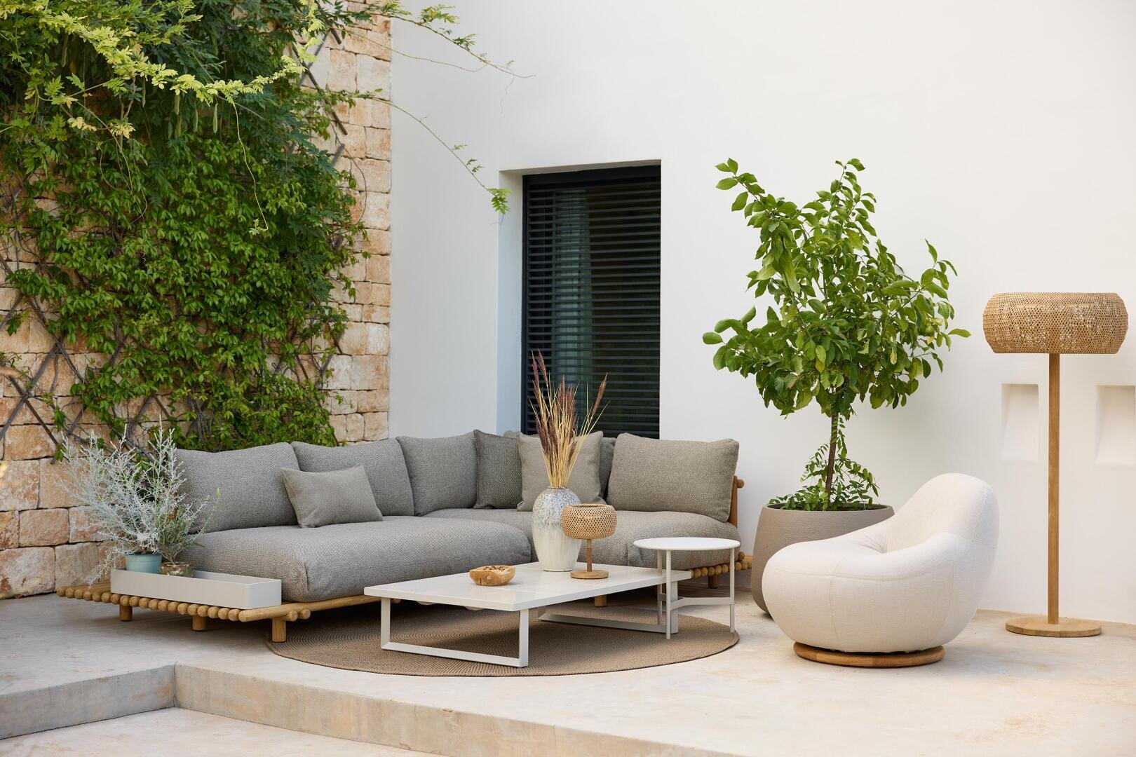 A stylish outdoor terrace with a gray sectional sofa, a white round chair, a white coffee table with decoration, potted plants, a woven floor lamp and stone and white walls with green vines.